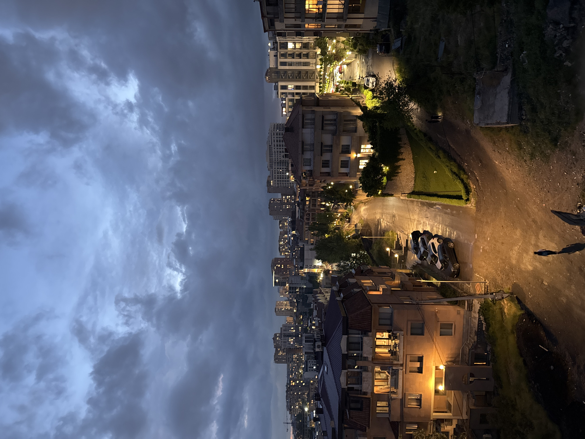 Captured from a high vantage point in Yerevan, Armenia, the scene portrays a twilight cityscape. The left side of the image is dominated by a heavy, cloudy sky. As the view descends, buildings densely populate the landscape, with numerous windows illuminated. A street cuts through the built environment, lit by streetlights and car headlights. Cars are parked along the roadside. The area appears to be a residential neighborhood. The right side of the image transitions into a dirt road adjacent to a grassy area. A human shadow is visible on the dirt road.