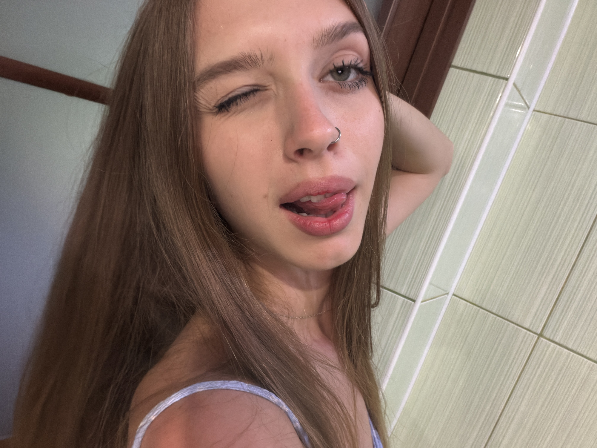 This media file is a close-up, self-portrait of a young woman with long, brown hair. She is looking directly at the camera with her left eye closed in a wink and her right eye open. Her tongue is playfully sticking out. She is wearing a thin necklace and a light-colored top with thin straps. A silver hoop is visible in her left nostril.
The background appears to be a bathroom, indicated by the tiled wall with vertical lines. The lighting suggests it could be daytime, or the interior lights are on. There is no visible text in the image. The overall mood is playful and suggestive.