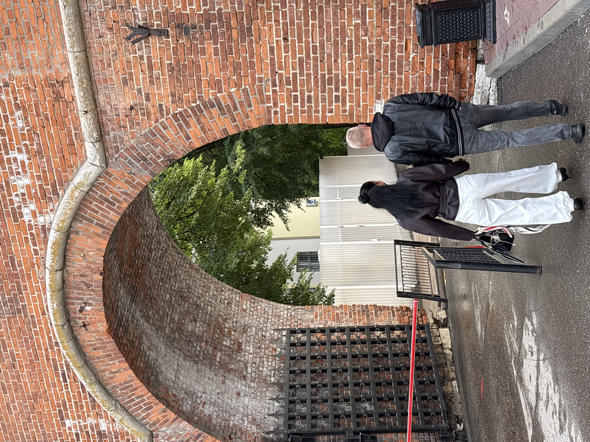 In Nizhny Novgorod, Russia, two individuals, a woman and a man, are walking hand-in-hand away from the camera, passing through a large arched brick gateway. The woman has long dark hair and is wearing a dark brown jacket and loose white pants. The man has short light-colored hair and is wearing a black hooded leather jacket and grey jeans. They are walking on a wet asphalt surface, suggesting it might be raining or has recently rained. To their left, a black metal gate with a decorative lattice pattern is visible, with a red barrier stretched across the path in front of it. The gateway itself is constructed from aged red brick, with a lighter colored stone trim lining the arch. Beyond the gateway, lush green trees are visible, along with a white fence and part of a building. To the right of the couple, near the brick wall, is a black ornamental trash receptacle. The scene appears to be a historical or old part of the city, possibly a fortress or an entrance to a historic site.