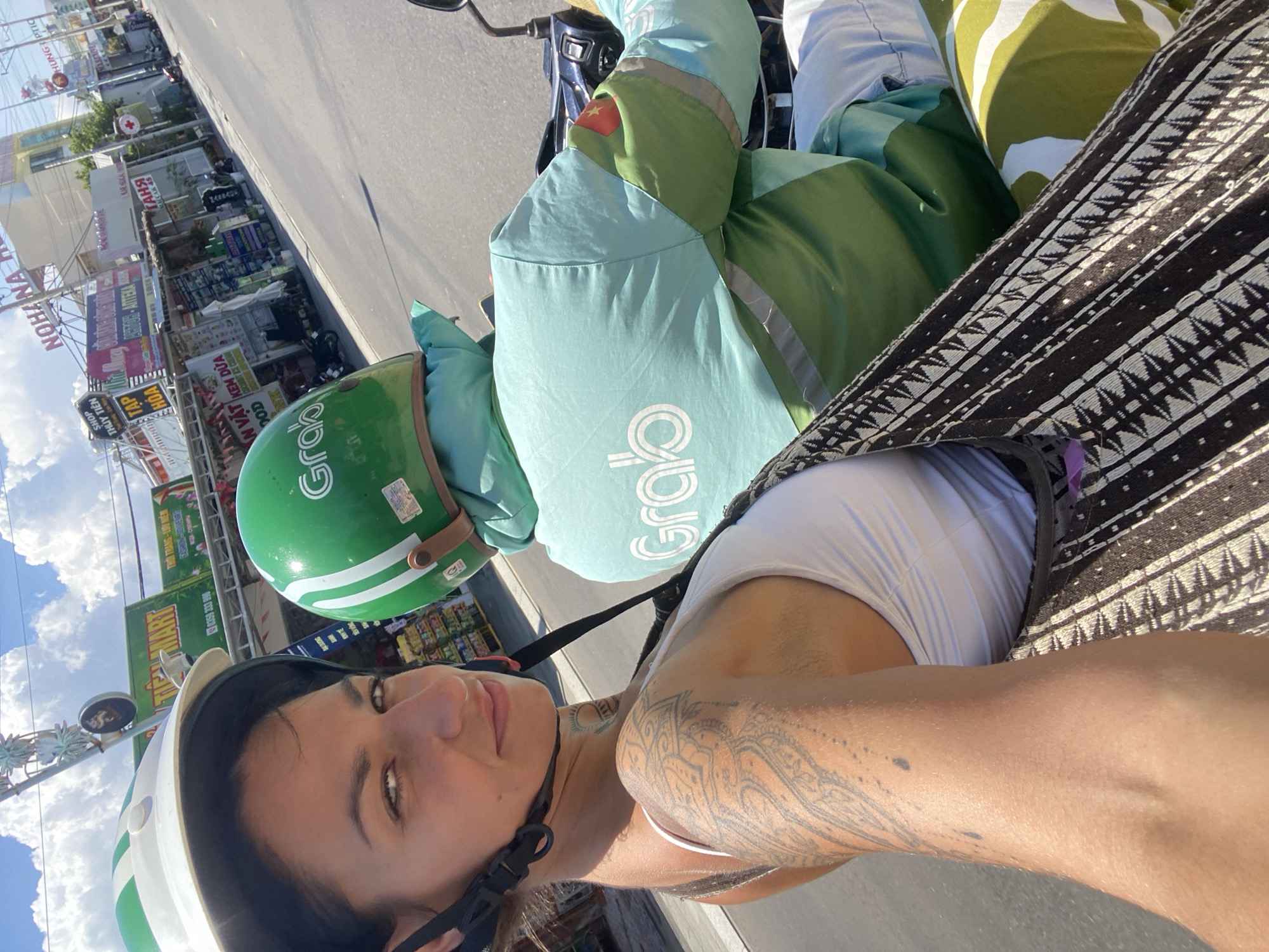 A woman with dark hair and tattoos on her arm and neck is riding on the back of a scooter. She is wearing a white tank top and a white and green helmet. The scooter driver, wearing a green Grab uniform and helmet, is in front of her. They are on a paved road in a city, with shops and buildings lining the street. Signs in Vietnamese are visible on the buildings. The sky is blue with some clouds. The location is Phu Thuy Ward, Vietnam.