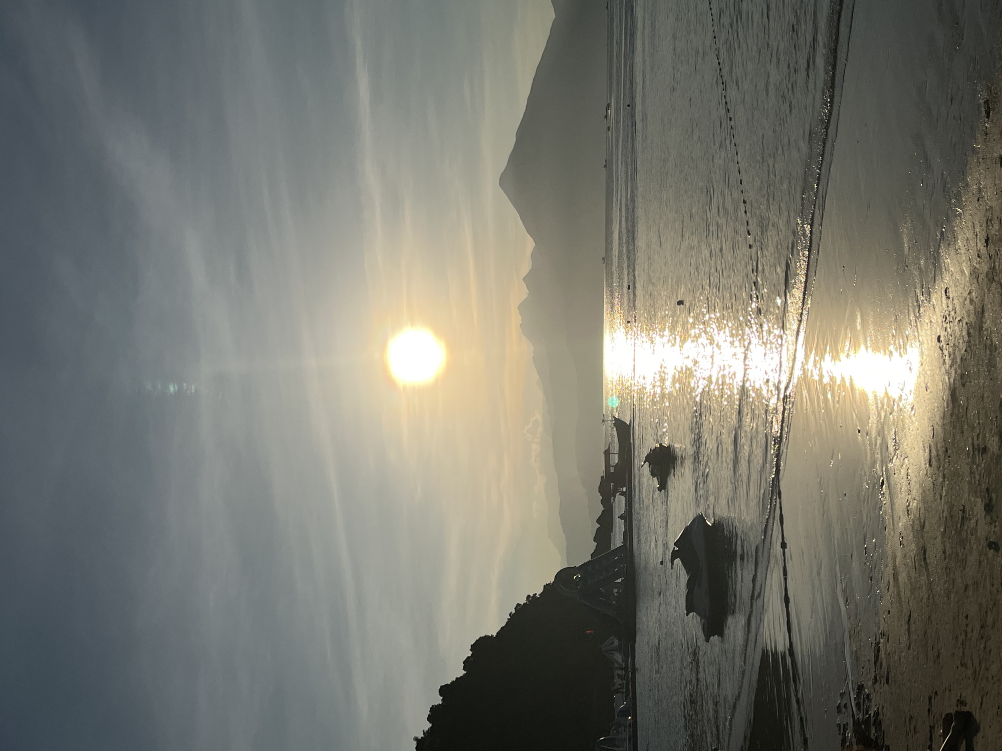 The image, captured in Đà Nẵng, Vietnam, depicts a serene coastal or lakeside landscape during golden hour, likely sunset. A bright, radiant sun is positioned prominently on the left side of the frame, casting a brilliant, shimmering path of light across the calm water towards the right. The intense sun creates a significant lens flare and imbues the entire scene with a warm, hazy glow.
The sky, visible in the upper portion, is partially obscured by soft, wispy clouds that diffuse the sunlight. In the mid-ground, a range of mountains or large hills appears in silhouette, their peaks subtly outlined against the diffused sky, contributing to the layered depth of the scene. The foreground on the lower left is anchored by a dark, tree-covered hill or shoreline, its features largely obscured by the backlighting.
Several small boats, possibly fishing vessels, are scattered across the water, particularly near the foreground landmass, appearing as dark silhouettes against the sun's reflection. The surface of the water reflects the sky's soft hues and the sun's intense sparkle, creating a dynamic texture with visible ripples. The overall atmosphere is tranquil and peaceful, highlighting the natural beauty of the environment at dusk or dawn. There is no visible text in the image.