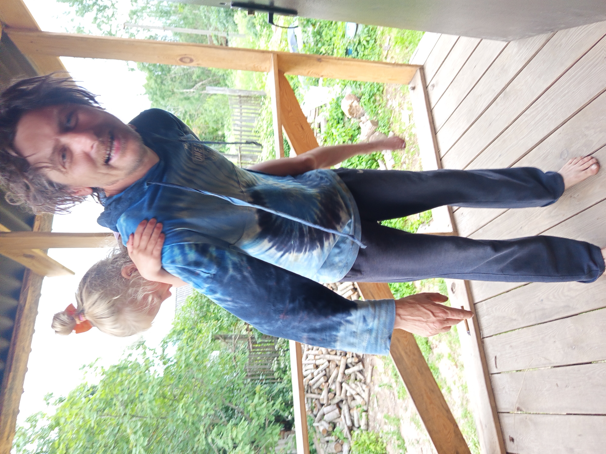 A man is giving a piggyback ride to a young child on a wooden porch in Ostrovtsy, Russia. The man, who is wearing a blue tie-dye hoodie and dark pants, is smiling and looking towards the camera. The child, who has blonde hair tied up with an orange bow, is facing away from the camera, looking down. In the background, there is a pile of firewood and a wooden fence, suggesting a rural or semi-rural setting. The porch appears to be attached to a wooden building. The lighting suggests it is daytime.