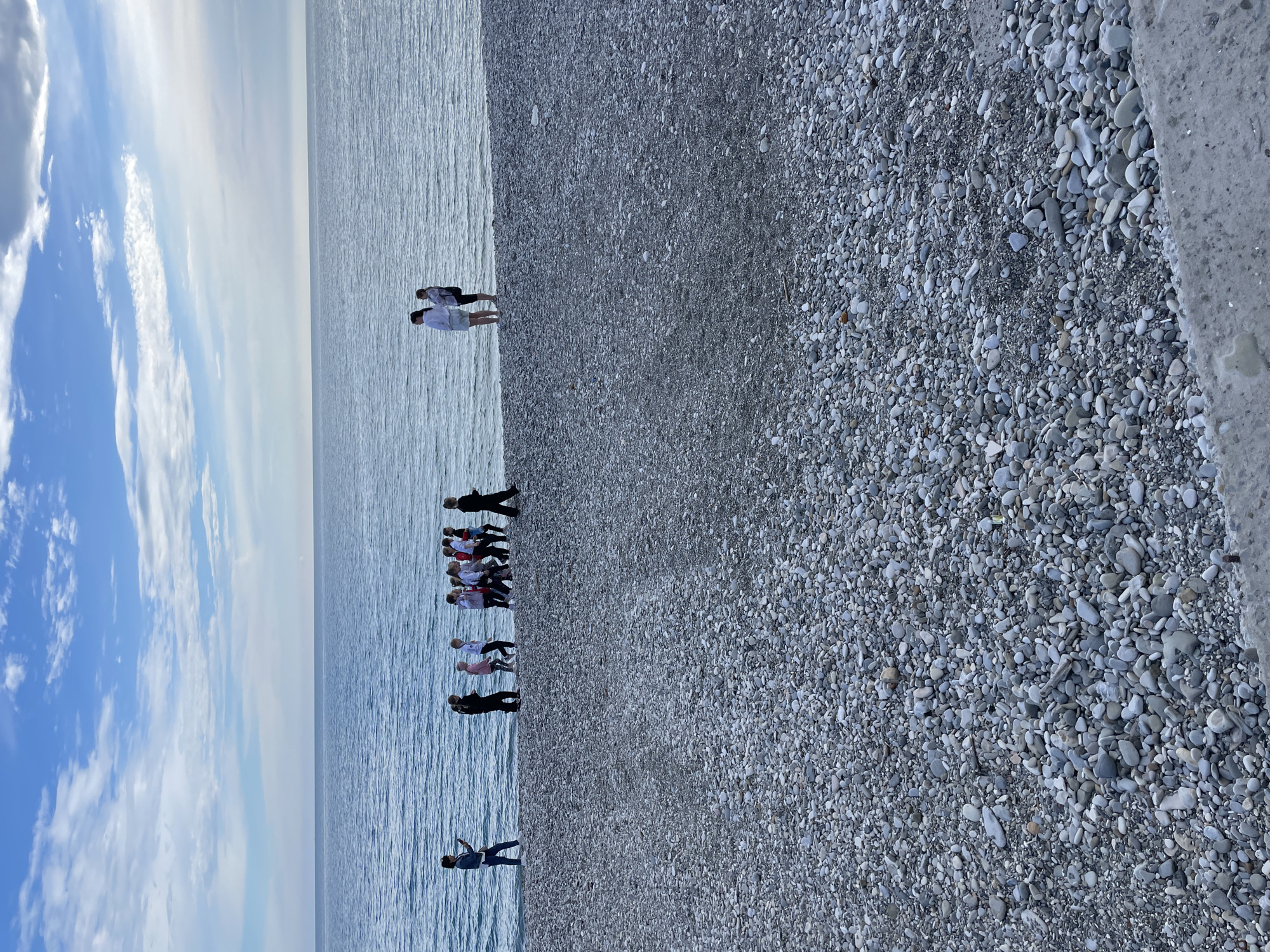 The image depicts a shoreline in Olginka, Russia. A pebble beach dominates the right side of the frame, with the sea occupying the left. People are scattered along the shoreline: a pair of individuals stand furthest away, closer to the center, there is a cluster of individuals in white shirts; other individuals stand more distantly. The sky features clouds, with the sun reflecting on the water.