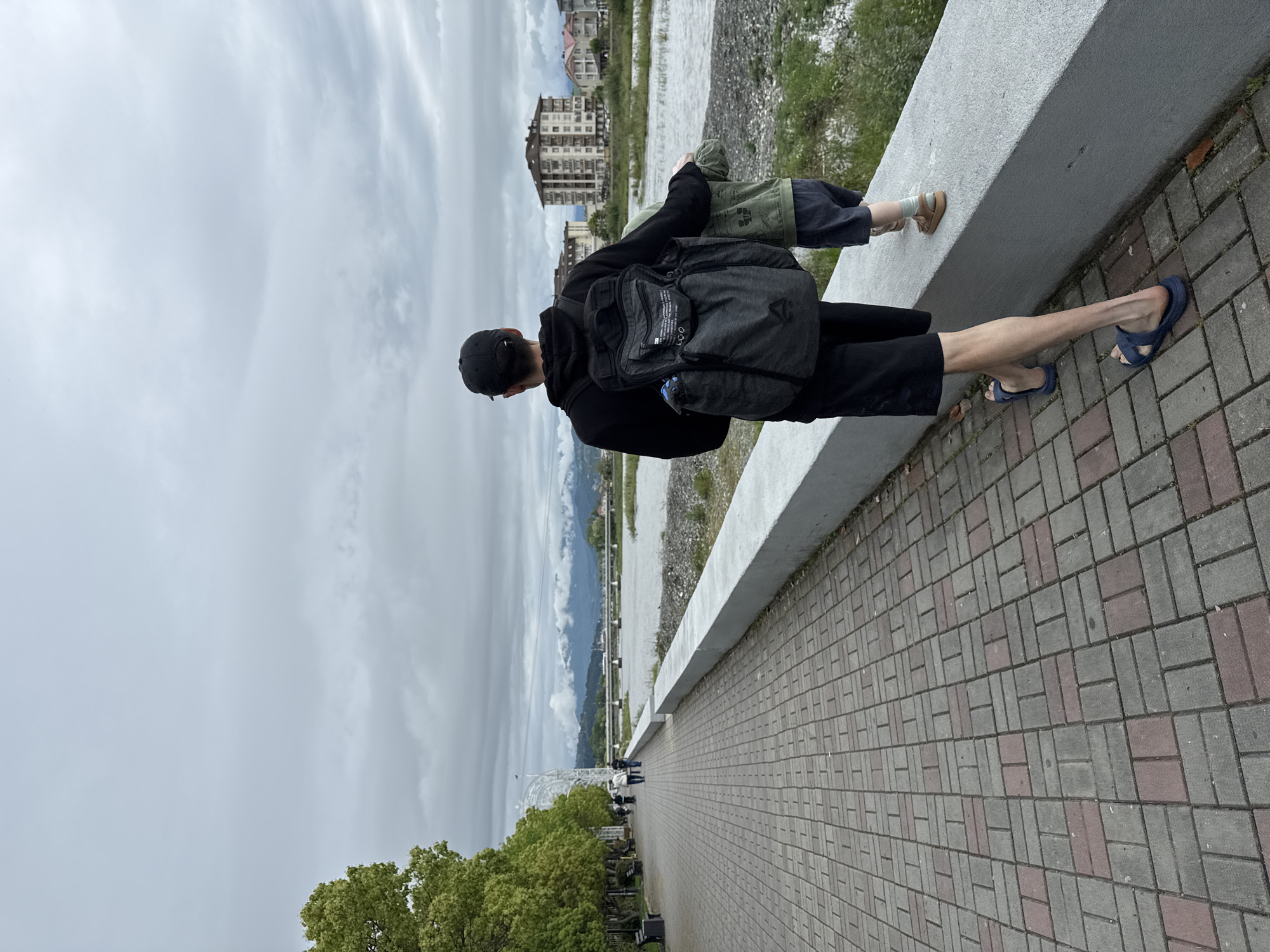 A man and a child stand on a paved walkway next to a low concrete wall, overlooking a river. The man is wearing a black hooded jacket, black shorts, a black baseball cap, and blue sandals. He has a black backpack on. The child is wearing a green jacket, dark-colored pants, and brown shoes. The child is leaning against the concrete wall. Behind them is a river with buildings on the opposite bank. In the background, there are mountains and a cloudy sky. The walkway is paved with grey and red bricks in a herringbone pattern. There is a row of trees along the walkway with benches. In the distance, other people can be seen walking along the path. The setting appears to be the city of Sirius, Russia, indicated by the modern buildings and riverside walkway.