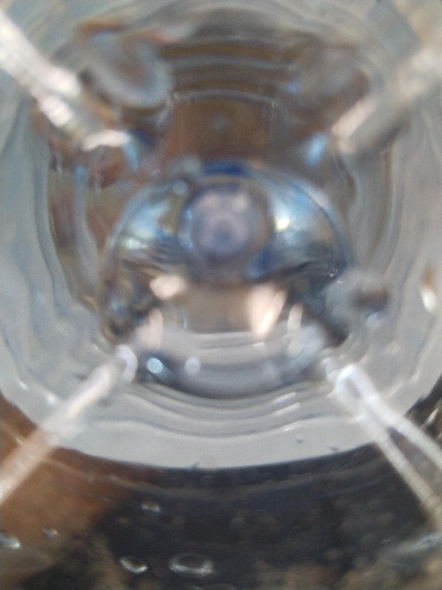 The image provides a close-up, top-down view into a clear plastic bottle, likely filled with water, given the reflections and the presence of small air bubbles. The focus is on the interior of the bottle, with the plastic walls creating a circular frame. Light refracts through the water, creating distorted shapes and reflections of the surrounding environment, which appears to be indoors with warm lighting. The lower portion of the image shows a dark, textured surface with more prominent bubbles, suggesting it might be the bottom of the bottle or a surface beneath it.
There are no discernible people, animals, or specific objects within the bottle's contents that can be clearly identified due to the abstract and distorted nature of the view. The scene is abstract, focusing on light, water, and the plastic material of the bottle.
There is no activity or event taking place, as the image captures a static view. The notable details are the optical effects of light and water, the texture of the plastic, and the presence of air bubbles. No specific time of day, weather, or emotions are conveyed. No text is visible in the image. The provided location context, "Гожскі сельскі Савет, Belarus," indicates a geographical setting, but this context is not visually represented in the image itself.