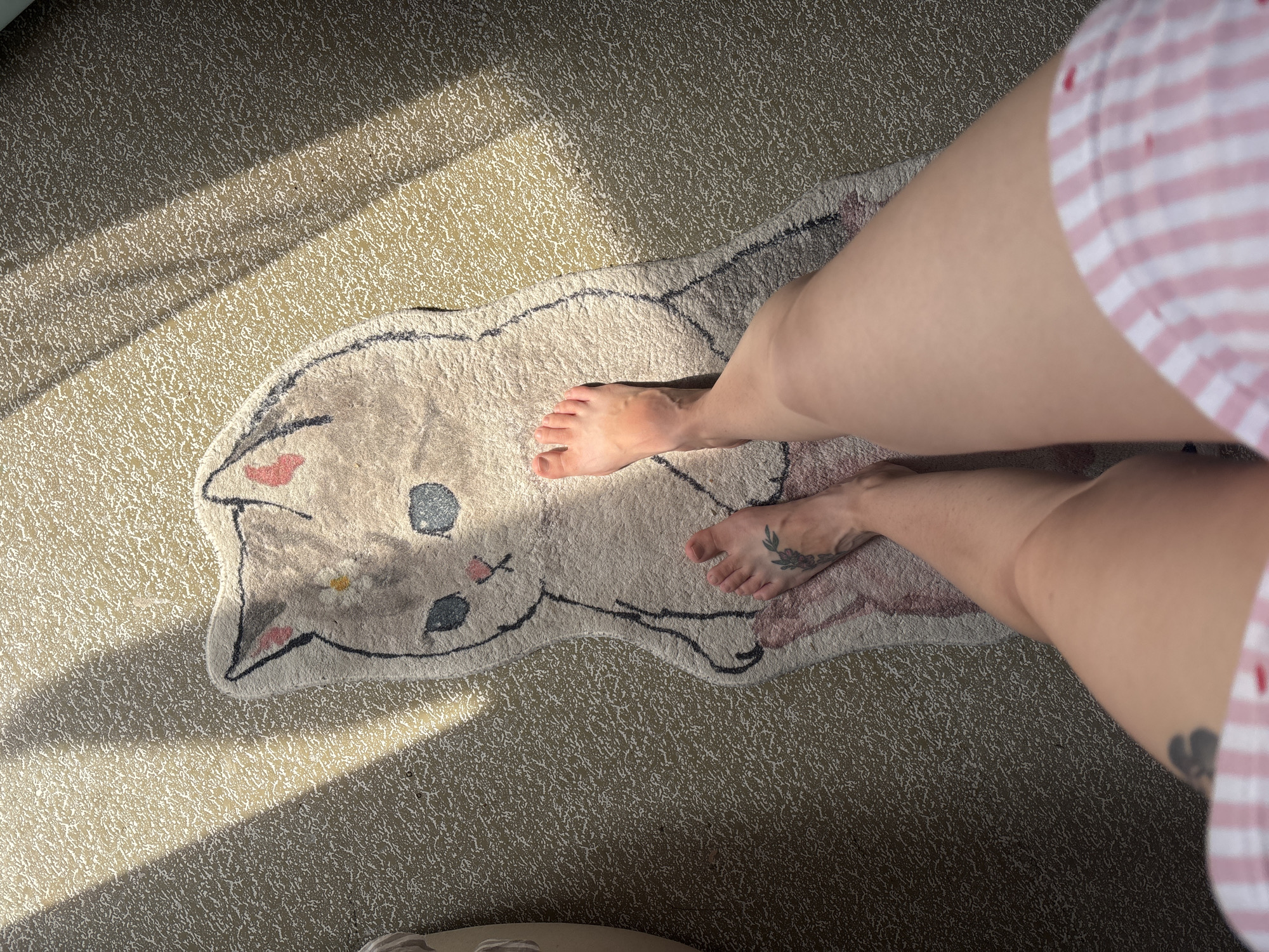The image shows a person's bare legs and feet standing on a rug shaped like a cat. The rug is light brown with black outlines and has a cartoonish cat face. The person's feet have tattoos on them. The environment appears to be indoors, with sunlight casting shadows on the floor. The scene is not located in Naberezhnye Chelny, Russia.
