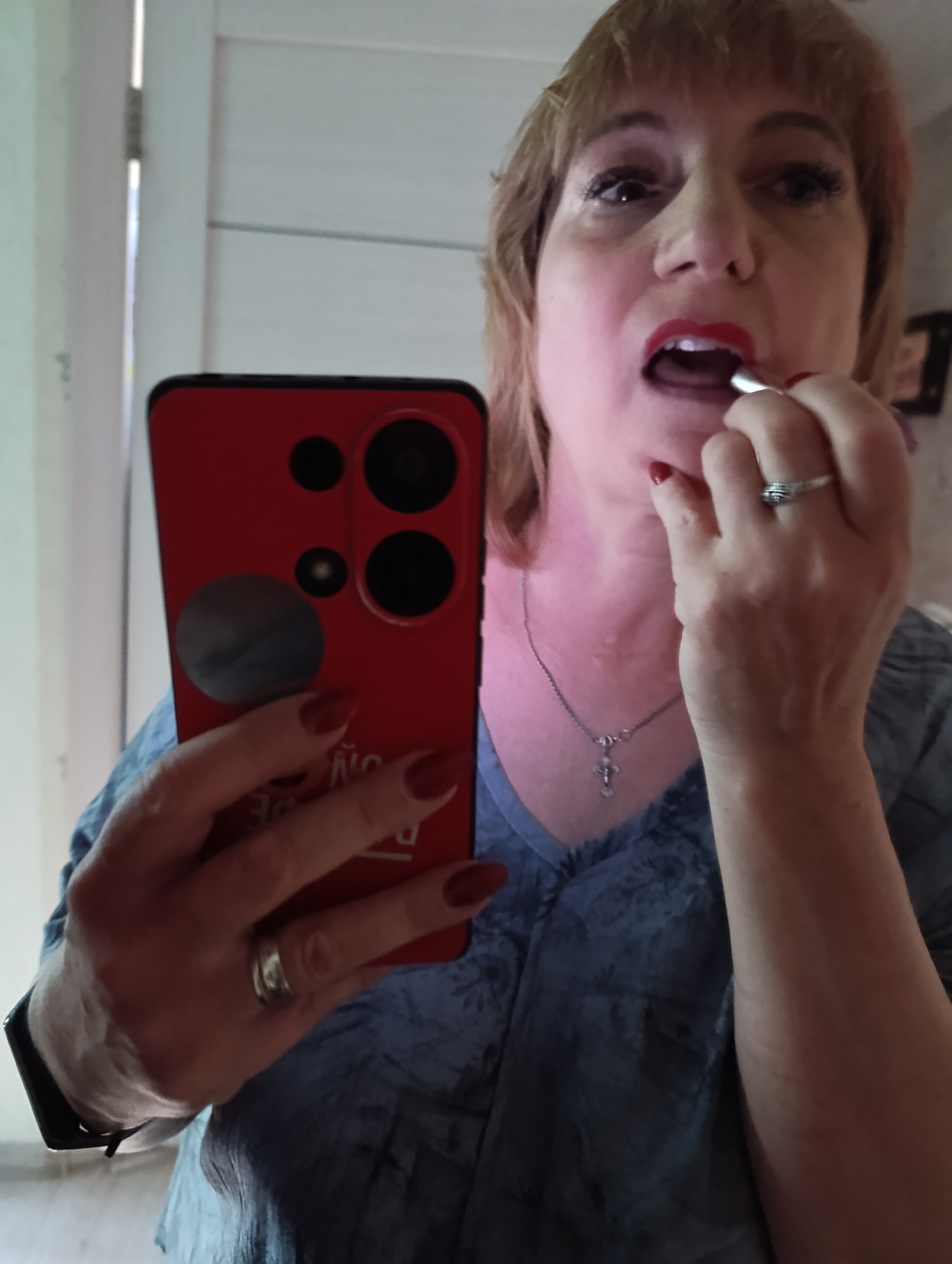 A medium shot captures a woman applying red lipstick to her lips while holding a red-cased smartphone in her left hand, taking a selfie. The woman, with blonde hair and light skin, is wearing a blue V-neck top. She has long, red-painted nails. A silver necklace with a cross pendant rests on her neck. Her right hand is raised, holding the lipstick to her lips. The smartphone is positioned to reflect her face. The background appears to be an indoor setting with a light-colored wall and vertical paneling, possibly in a home or public restroom. The image provides no specific information about the location being Tugulym, Russia.