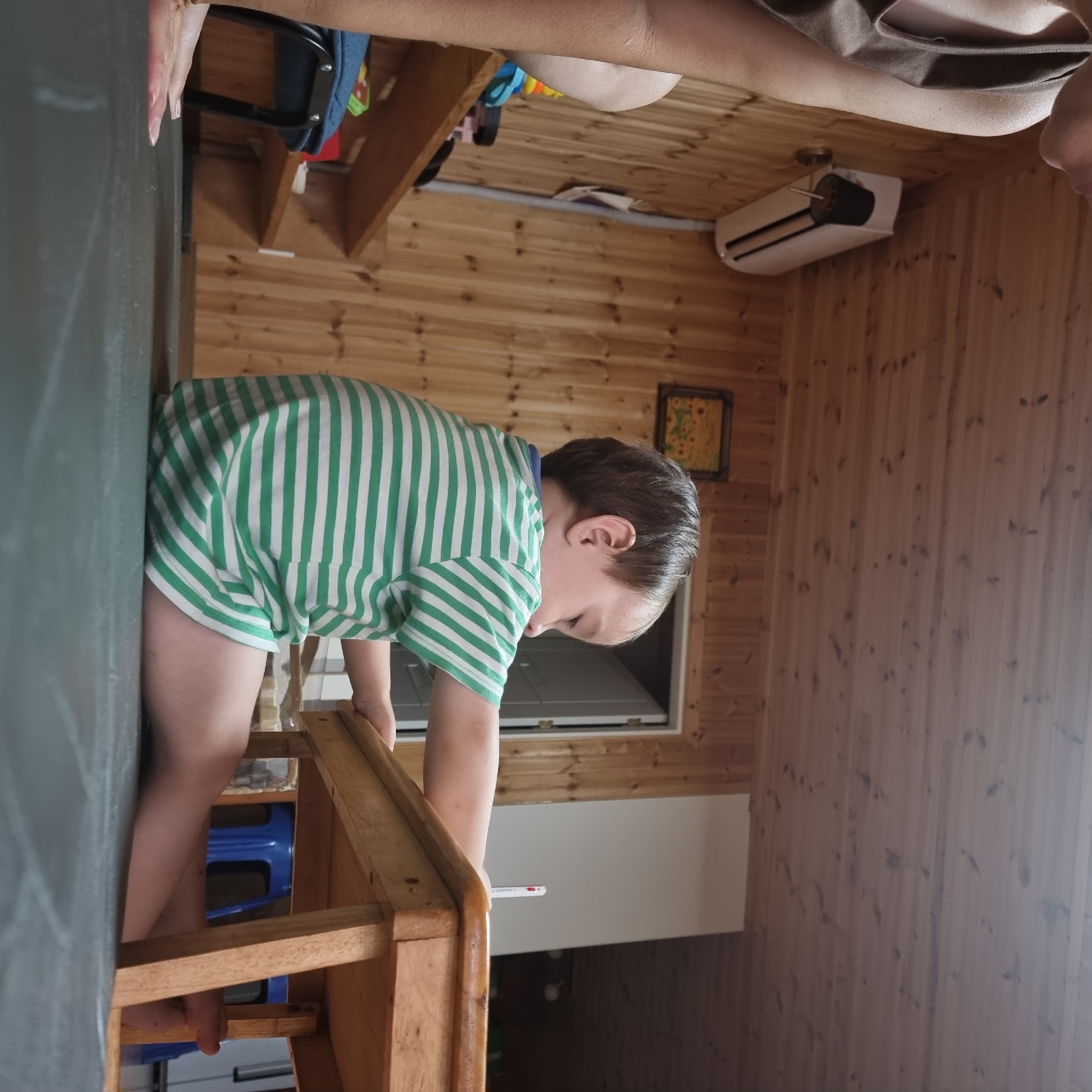 The image depicts a young child wearing a green and white striped t-shirt, sitting on a dark grey floor in a room with wood-paneled walls. The child is looking down and appears to be interacting with a small wooden table, on which a white object with a red tip is visible, possibly a pen or crayon. To the left of the child, a portion of an adult's arm and shoulder, wearing a brown garment, is visible. In the background, there is a white air conditioning unit mounted on the wall, a framed picture of sunflowers, and a partially open doorway. Further in the background, a blue plastic stool is partially visible. The environment suggests an indoor domestic setting. The image was taken in Dunnae-myeon, South Korea.