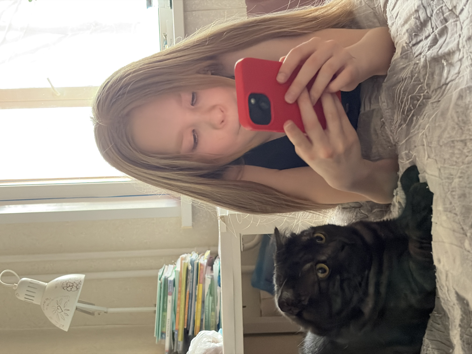 A young girl with long blonde hair is lying on a bed, holding a red smartphone, possibly taking a photo. A black cat with wide, yellow eyes is looking towards the camera from below the girl, partially obscured by the bed cover. In the background is a window, a white lamp, and a shelf with books. The location, Izhevsk, Russia, cannot be confirmed based on the image content alone.