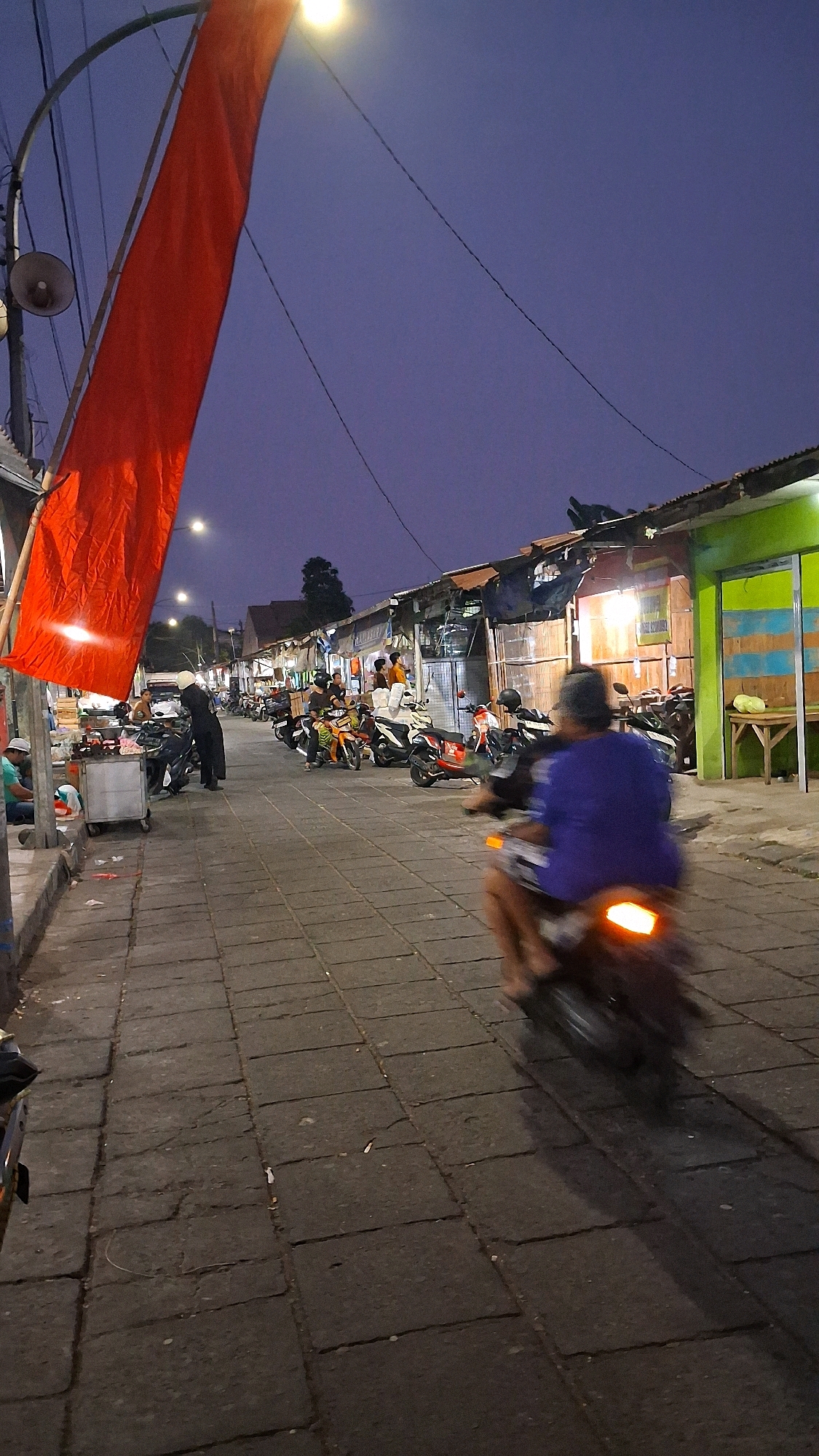 The image depicts a street scene in a market area at dusk, possibly in Yogyakarta, Indonesia, based on the style of the stalls and the presence of motorcycles. A large, bright red flag or banner hangs on the left side of the frame, partially obscuring a street light. The street is paved with rectangular stone tiles.
On the right side of the street, a row of market stalls or shops lines the path. These structures have corrugated metal roofs and wooden or bamboo walls, some painted in vibrant colors like green and yellow. Lights are visible inside some of the stalls, indicating activity.
Several motorcycles are parked along the side of the street, suggesting this is a common mode of transportation. A person is riding a motorcycle in the foreground, moving away from the camera. The rider is wearing a blue shirt, and the motorcycle's taillight is illuminated, creating a blur effect due to motion. Other people can be seen in the background, some standing near the stalls and others near parked motorcycles. One individual in the mid-ground is wearing a dark outfit and a white helmet, standing near a food cart.
The sky is a deep twilight blue. The overall impression is of a bustling market transitioning from day to night, with a mix of commercial activity and local life.