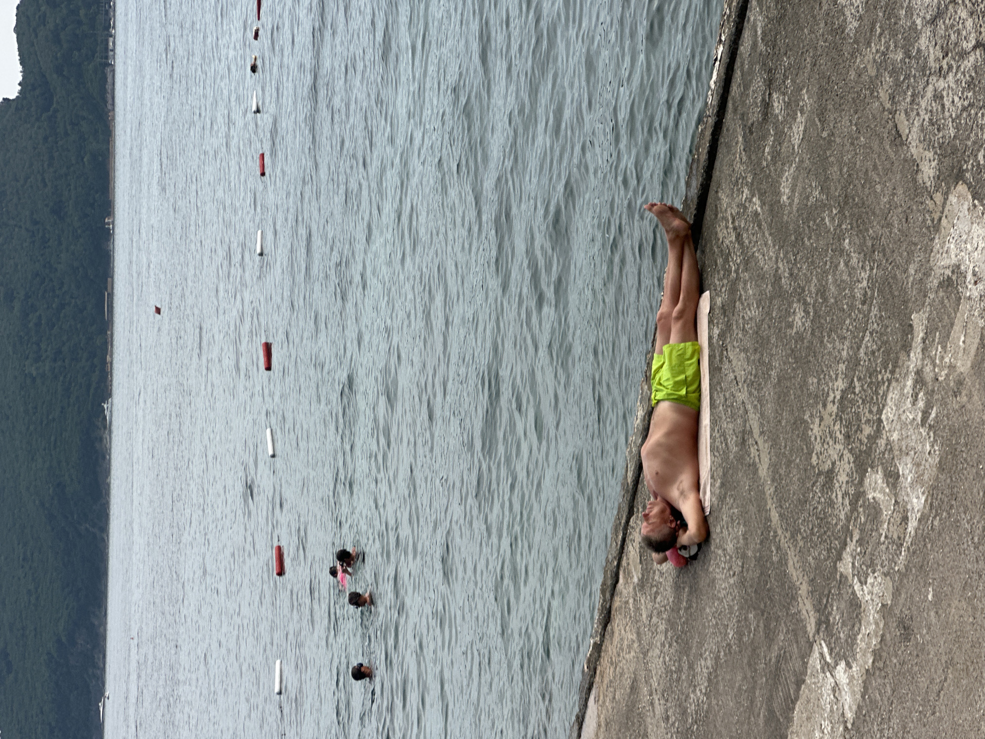 In Budva, Montenegro, on a cloudy day, a man lies on his back on a concrete embankment, sunbathing on a towel. He is wearing bright green swim trunks and has his arms behind his head. His eyes are closed, and he appears to be sleeping or resting. The embankment is a rough, gray surface with some debris scattered on it. To the right, the sea stretches out, its surface rippling. In the distance, a line of buoys marks a swimming area. Further out, several people are swimming and playing in the water. Behind the sea, a lush green hillside rises, partially obscuring buildings and trees along its base. The overall scene depicts a tranquil moment of leisure by the sea.