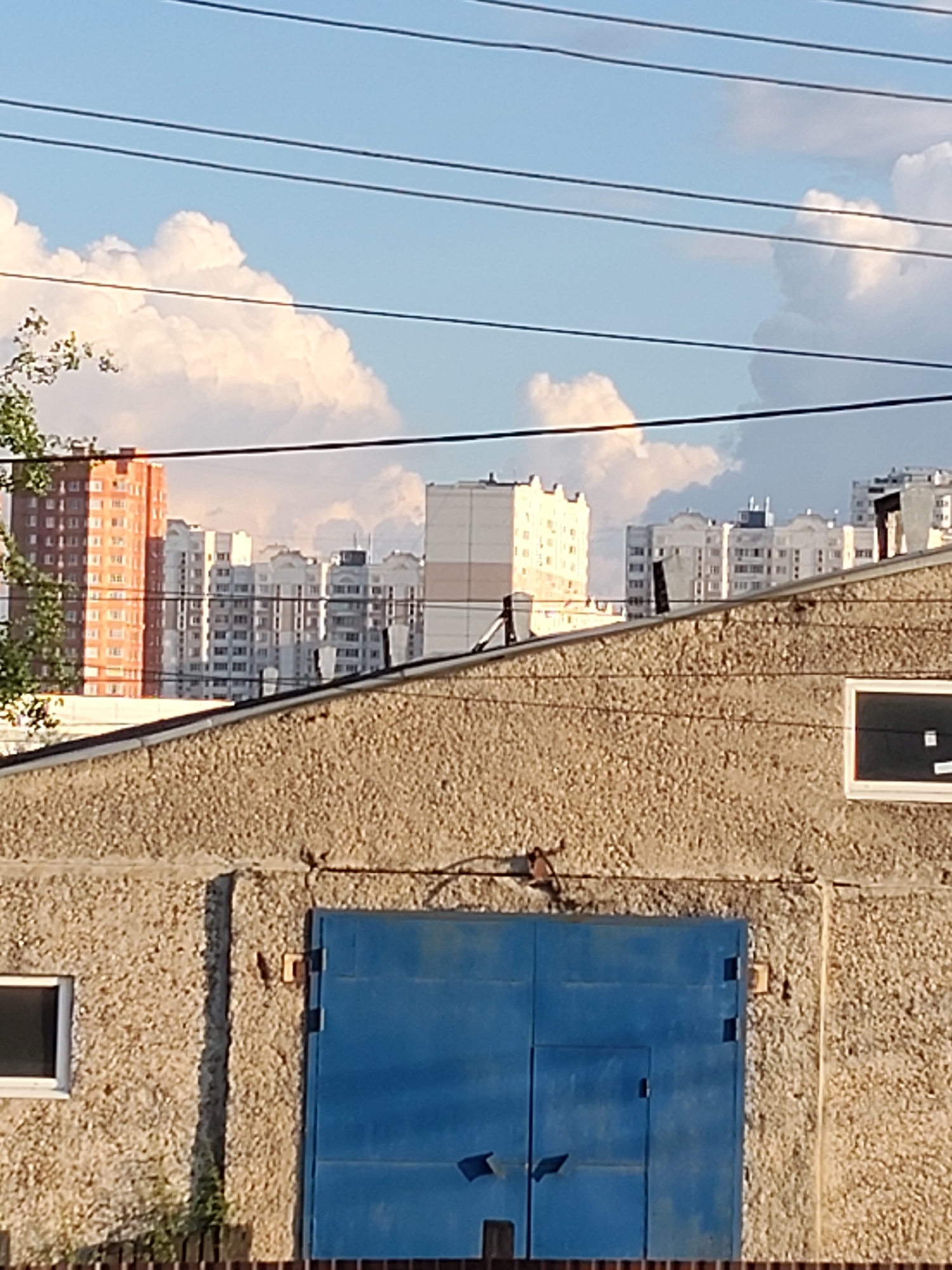 The image shows a view of apartment buildings and utility wires against a cloudy sky. In the foreground, a textured concrete building with a large blue garage door and a small window is visible. In the middle ground, several multi-story apartment buildings are seen, some with orange facades and others with white and grey facades. The sky is filled with large, white, fluffy clouds. The scene is likely a residential area in Chekhov, Russia, given the architectural style of the buildings and the presence of utility wires, which are common in Russian cities.