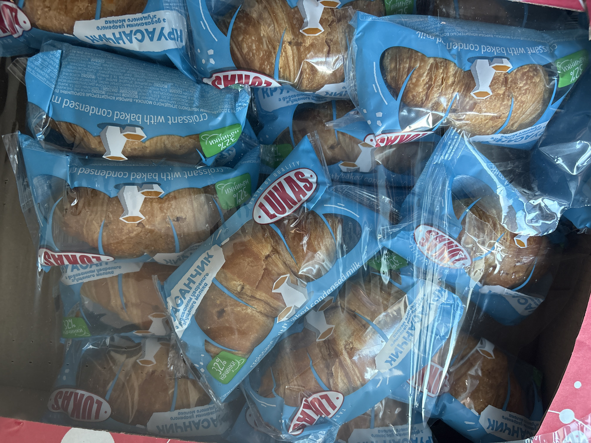 The image shows a close-up, overhead view of a cardboard box filled with individually packaged croissants. The croissants are golden brown and appear to be of the crescent shape. Each croissant is enclosed in a clear plastic wrapper with a blue and white design. Prominently featured on the packaging is the brand name "LUKAS" in red lettering within an oval. There are also labels in Ukrainian, some of which indicate "croissant with baked condensed milk" and "32% filling." The packaging also features a stylized illustration of a white cow with yellow horns, possibly symbolizing milk. The croissants are tightly packed in the box, obscuring a clear view of the entire contents. The overall impression is of a retail product ready for sale or transport. The image does not provide any specific information about the city of Dnipro, Ukraine, or any details about people, actions, or interactions.