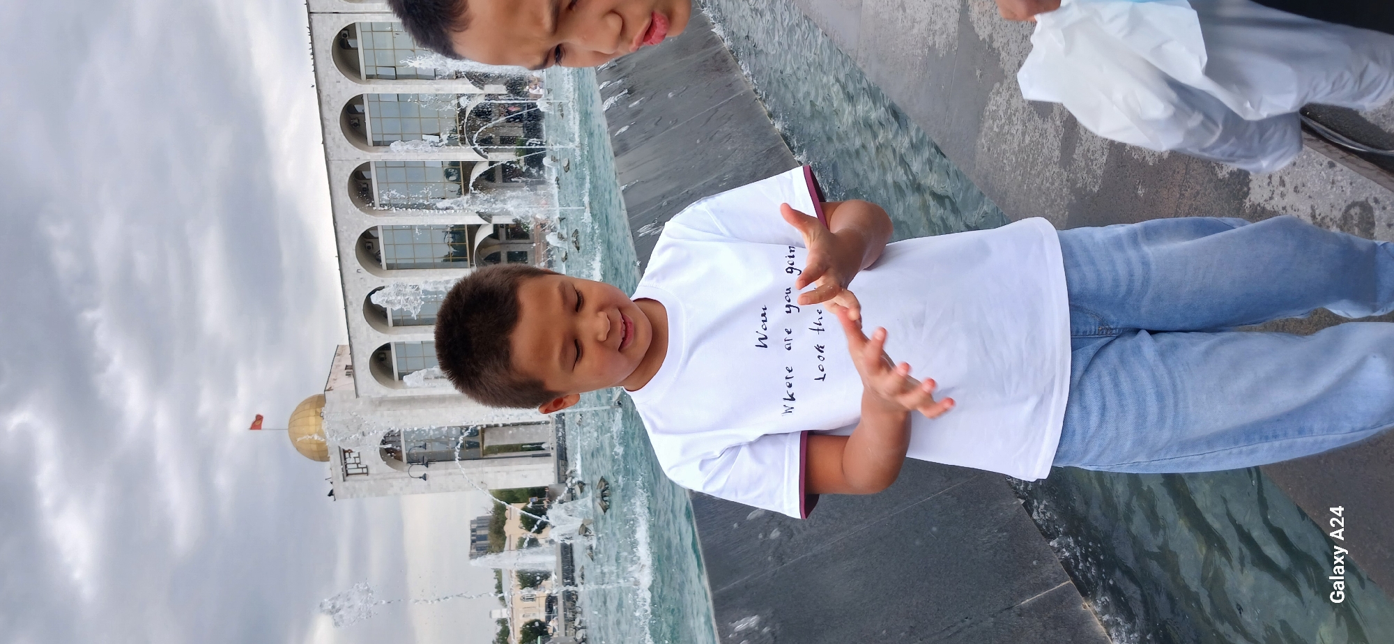 In Bishkek, Kyrgyzstan, in front of the State Historical Museum, two young boys are captured in a photograph near a large fountain. The boy on the left, wearing a white t-shirt with text that reads "Wow, Where are you going? Look the..." and blue denim jeans, is looking down at his hands, which are held in front of him as if interacting with something unseen. The boy on the right is partially visible, holding a white plastic bag. The background features the grand architecture of the museum with its distinctive arches and a golden dome topped by a flag. The fountain in the foreground is active, with water spraying upwards, reflecting the overcast sky.
