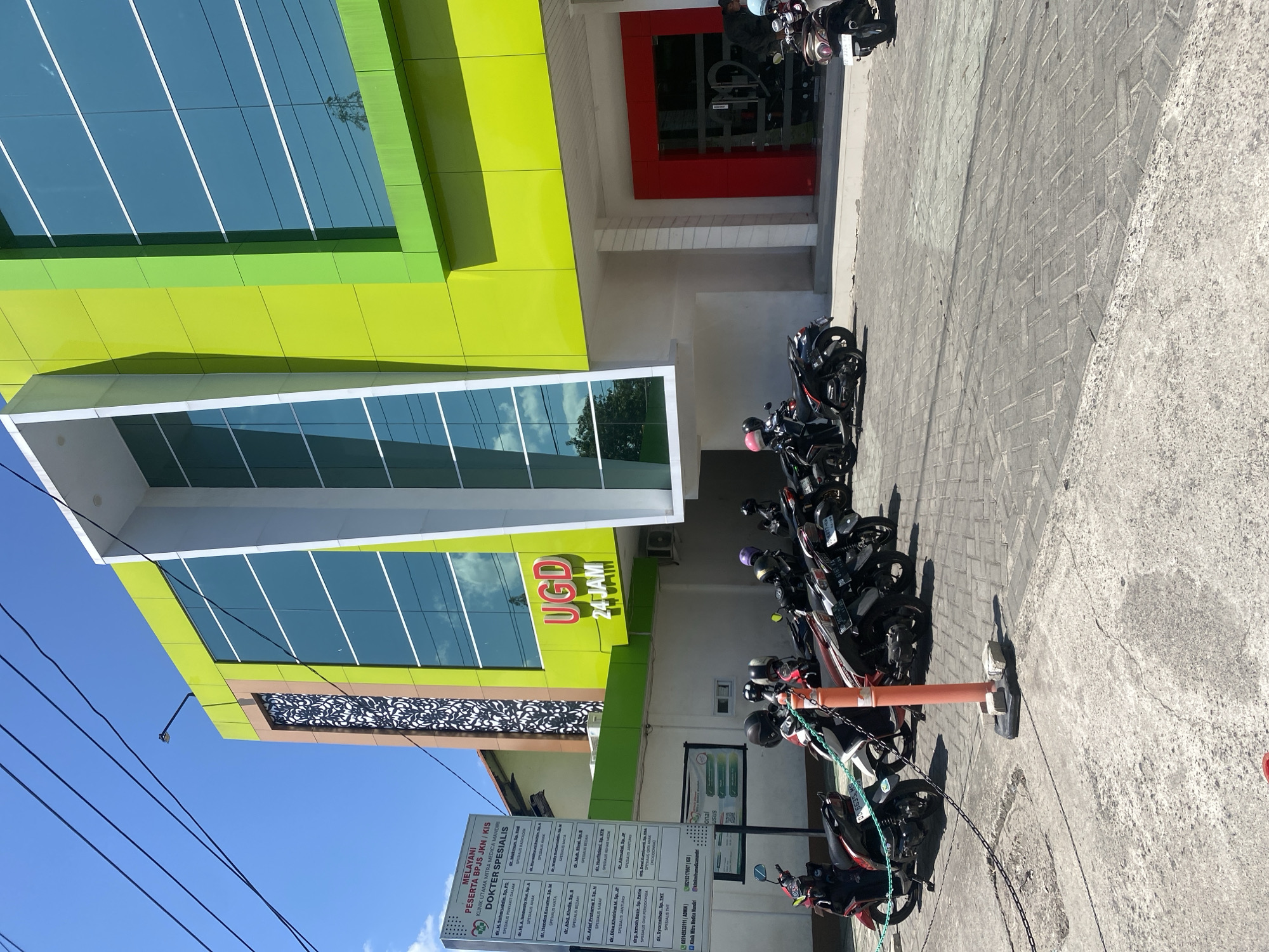 A modern, multi-story building with a bright green and lime green facade stands under a clear blue sky in Bantaeng, Indonesia. The building features large, reflective windows and a prominent entrance with a bright red door. Above the entrance, a sign reads "UGD 24 JAM," indicating an emergency unit operating around the clock. In front of the building, a row of parked motorcycles, mostly scooters, are visible. Some are covered with chains. A few people are seen near the entrance, dressed casually. Power lines crisscross the sky above the building. The ground is paved with interlocking gray bricks. A white sign with text and a logo is posted to the left of the building.
