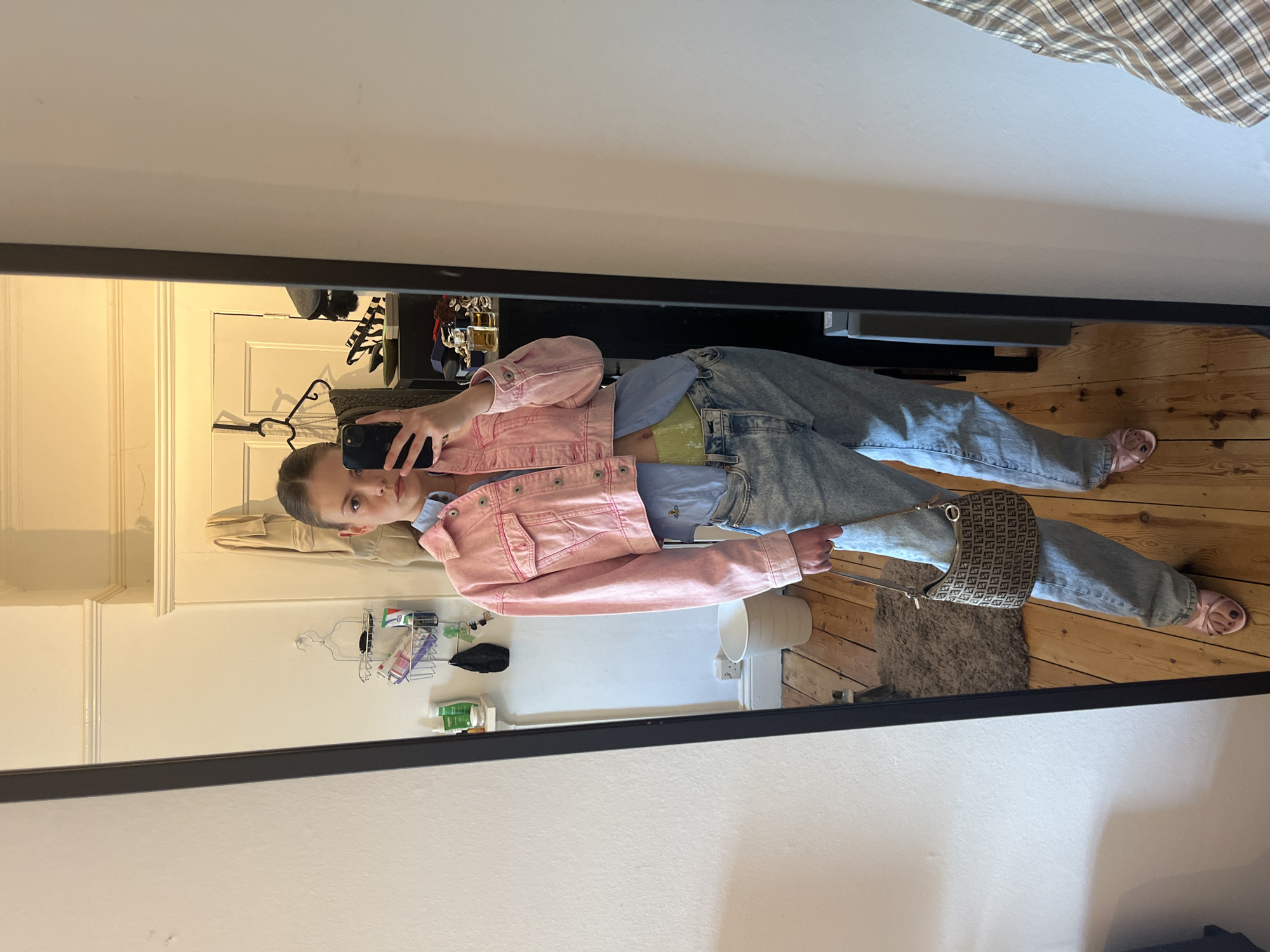 A young person takes a photo of themselves in a mirror. They stand on a wooden floor in what appears to be a bedroom. They are wearing light blue jeans, a light blue button-down shirt, a short pink denim jacket, and light pink slippers. They hold a smartphone in their left hand to take the picture. A purse hangs from their right shoulder. The mirror reflects the room behind them, showing a white wall, a shelf with various items, and a door. The location is implied to be in London, United Kingdom, but there are no specific landmarks or indicators visible in the image.