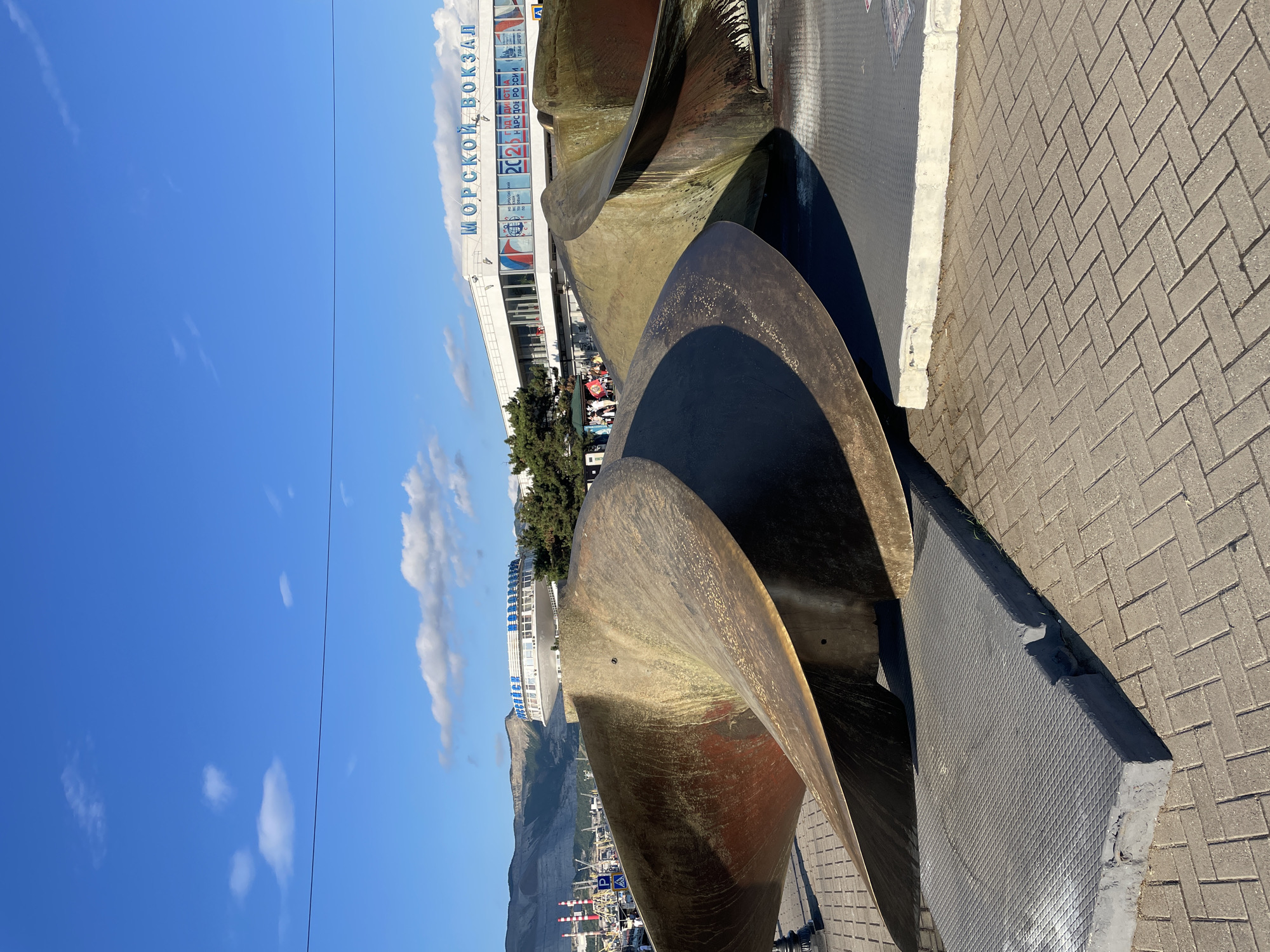 This is a photograph of a large metal sculpture resembling a ship's propeller, located in the city of Novorossiysk, Russia. The sculpture is situated outdoors on a paved walkway, with a building labeled "МОРСКОЙ ВОКЗАЛ" (Maritime Station) in the background. Mountains and industrial structures, likely part of the port, are visible in the distance. The sky is clear blue with some scattered clouds. There are a few people walking in the background, near the Maritime Station. The sculpture itself is weathered and shows signs of age, with a reddish-brown patina.