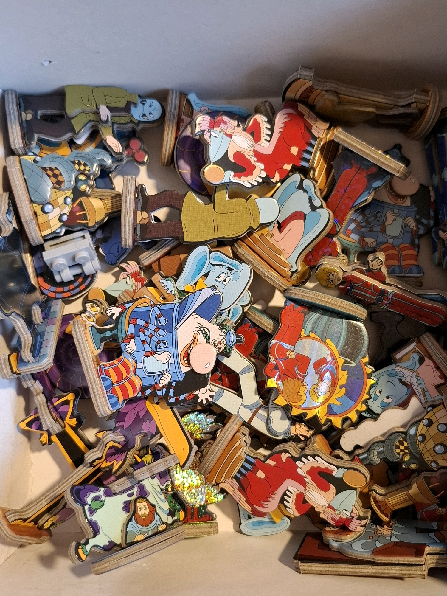 A box is filled with a jumble of puzzle pieces. The pieces are wooden, with colorful cartoon illustrations on them. The illustrations depict characters in various outfits, some appearing to be from a fantasy or science fiction genre. There are figures with blue skin, red outfits, and mechanical elements. The pieces are layered on top of each other, making it difficult to discern individual puzzles or their complete shapes. The overall impression is a chaotic collection of disassembled puzzle elements. The image is taken from a top-down perspective. This image has no relation to the city of Tyumen, Russia.