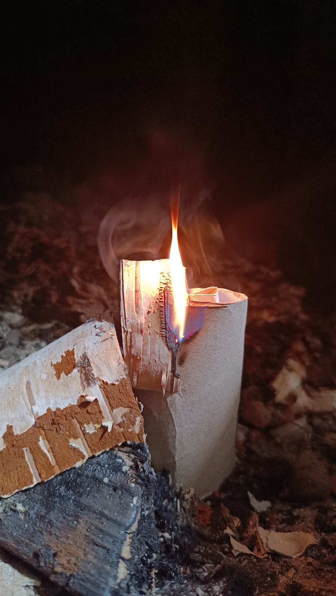 A close-up, eye-level shot shows a burning toilet paper tube on a dark, cluttered surface. The tube is upright and has a flickering flame emerging from its top, with wisps of smoke curling upwards and to the left. The left side of the tube is partially peeled back, revealing a rough, wood-like texture. To the left of the tube is a piece of wood with peeling bark and exposed inner material. The background is dark and indistinct, with hints of rough textures, possibly ash or debris. The scene is dimly lit, with the flame providing the primary light source, casting an orange glow on the surrounding objects.