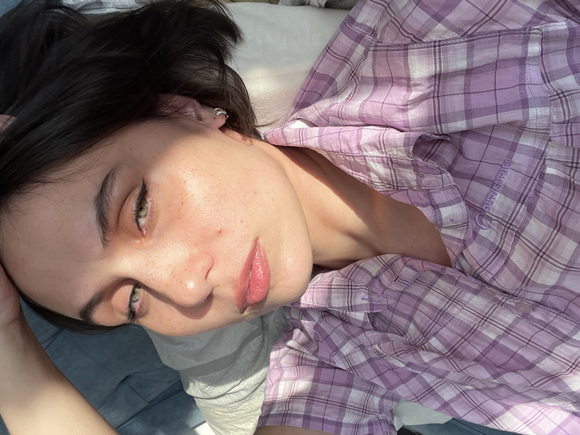 A close-up, high-angle shot captures a person with dark hair and striking green eyes, presumably in Izhevsk, Russia. The individual is lying down, looking towards the camera with a calm and serene expression. Their head is tilted, and natural light, likely from a window, casts strong highlights on their face, shoulder, and part of their purple and white plaid shirt, creating distinct shadows. The sunlight suggests it is either morning or late afternoon.
The person has visible freckles on their nose and cheeks, and subtle eyeliner defines their eyes. Their lips are painted a muted pink. A small, silver hoop earring is visible in their right ear. They are wearing a button-down shirt with a plaid pattern of varying shades of purple and white. Part of the shirt, specifically the word "SURVENTURE," is visible on the fabric, distorted slightly by the folds. The background appears to be an indoor setting, possibly a bed or couch, covered with light-colored fabric, contributing to a relaxed and intimate atmosphere.