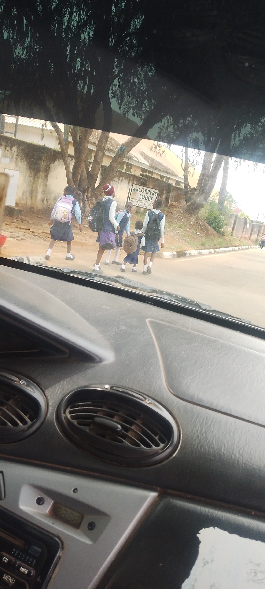 A group of five schoolchildren, four girls and one younger child, walk across a street in Jos, Nigeria. They are wearing matching school uniforms of blue shirts and dark blue/purple skirts. Three of the older girls have backpacks, and the younger child has a small brown backpack. The children appear to be crossing the street, with the oldest girl on the left looking back and smiling. A sign in the background reads "CORPERS LODGE". The scene is captured from inside a vehicle, with the dashboard visible in the foreground.