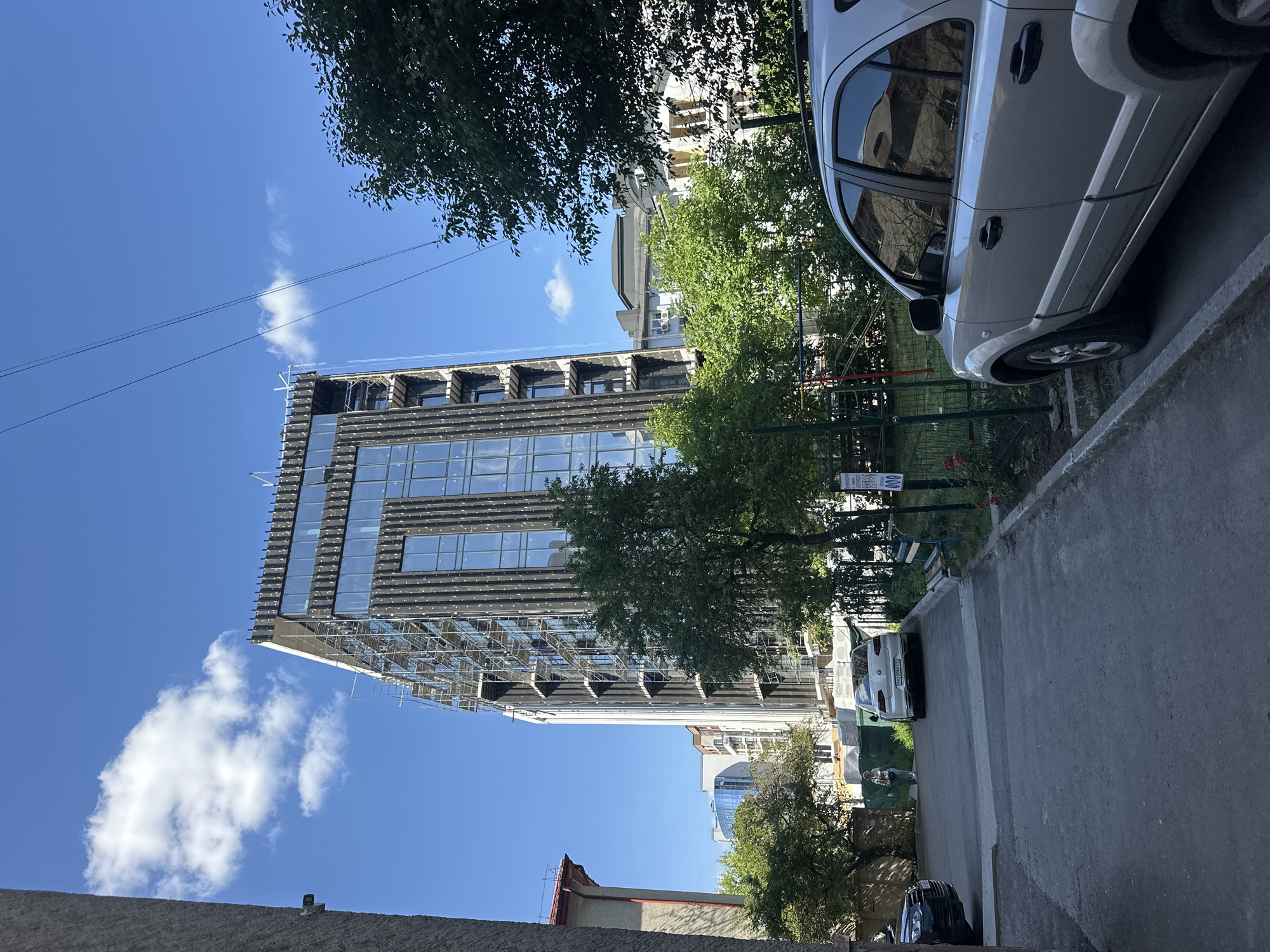 The image depicts an outdoor scene in Ternopil, Ukraine, on a clear, sunny day with scattered clouds. The perspective is from a street level, looking upwards and slightly to the side, with the image rotated 90 degrees clockwise.
The primary subjects are a modern, multi-story building undergoing construction or renovation, a parked silver SUV, and lush green trees. The building features a glass facade and scaffolding, suggesting ongoing work. The SUV is parked on the right side of the frame, close to a sidewalk and a grassy area with a fence. The trees are situated in front of the building and along the street, providing shade and greenery.
A person can be seen standing near the rear of the silver SUV. The time of day appears to be daytime, given the bright sunlight and the clear blue sky. The weather is pleasant and sunny.
Notable details include the scaffolding on the building, indicating active construction. The reflection in the SUV's windows shows parts of the surrounding buildings. Along the street, there is a paved surface and a concrete barrier.
There is some visible text on a sign near the parked SUV, but it is too blurry to be legible. The overall impression is one of an urban environment with a mix of older and newer architecture, and ongoing development. The scene is peaceful and orderly.