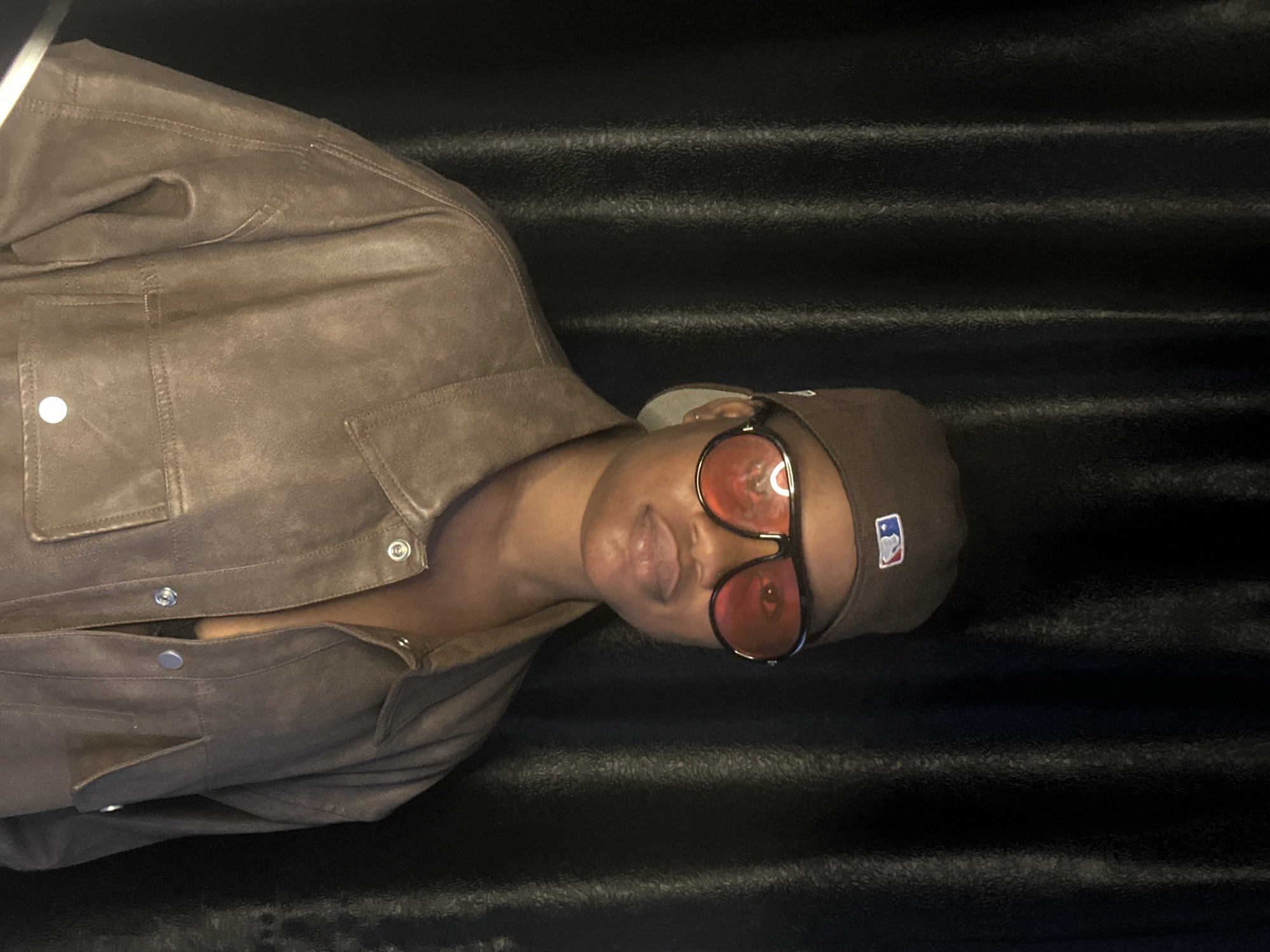 The image shows a person from the chest up, wearing a brown jacket and a brown baseball cap. The person is also wearing oversized sunglasses with red lenses. The background is dark and appears to be textured, with horizontal lines. The person's head is tilted back, and their mouth is slightly open. The overall lighting is dim, suggesting it might be nighttime or an indoor setting with low light. The MLB logo is visible on the baseball cap. The image is oriented vertically, with the person's head at the bottom and their shoulders at the top. The setting does not provide specific visual cues for the location of Badagry, Nigeria, beyond the provided context. No specific activity or event is clearly depicted, beyond the person posing for a picture. The emotion conveyed is ambiguous, possibly relaxed or playful due to the sunglasses and slight smile.