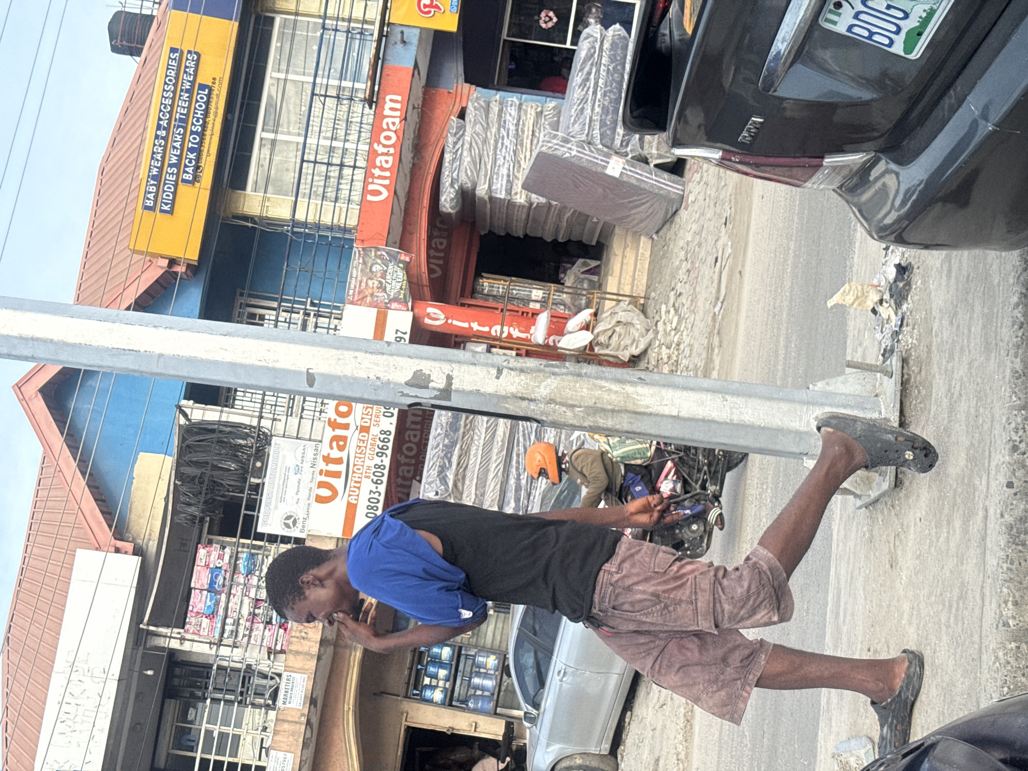A street scene in Ado, Nigeria, featuring a Black male wearing a black t-shirt, brown shorts, and black clogs. He is captured mid-stride on the sidewalk.
Background elements:
* Storefronts with signs reading "Vitafoam" and "BABY WEARS & ACCESSORIES...BACK TO SCHOOL."
* A black car partially visible on the right side of the frame with a license plate marked "BDG."
* Another car is seen parked behind the man.
* Other pedestrians and a motorcycle can be observed nearby.