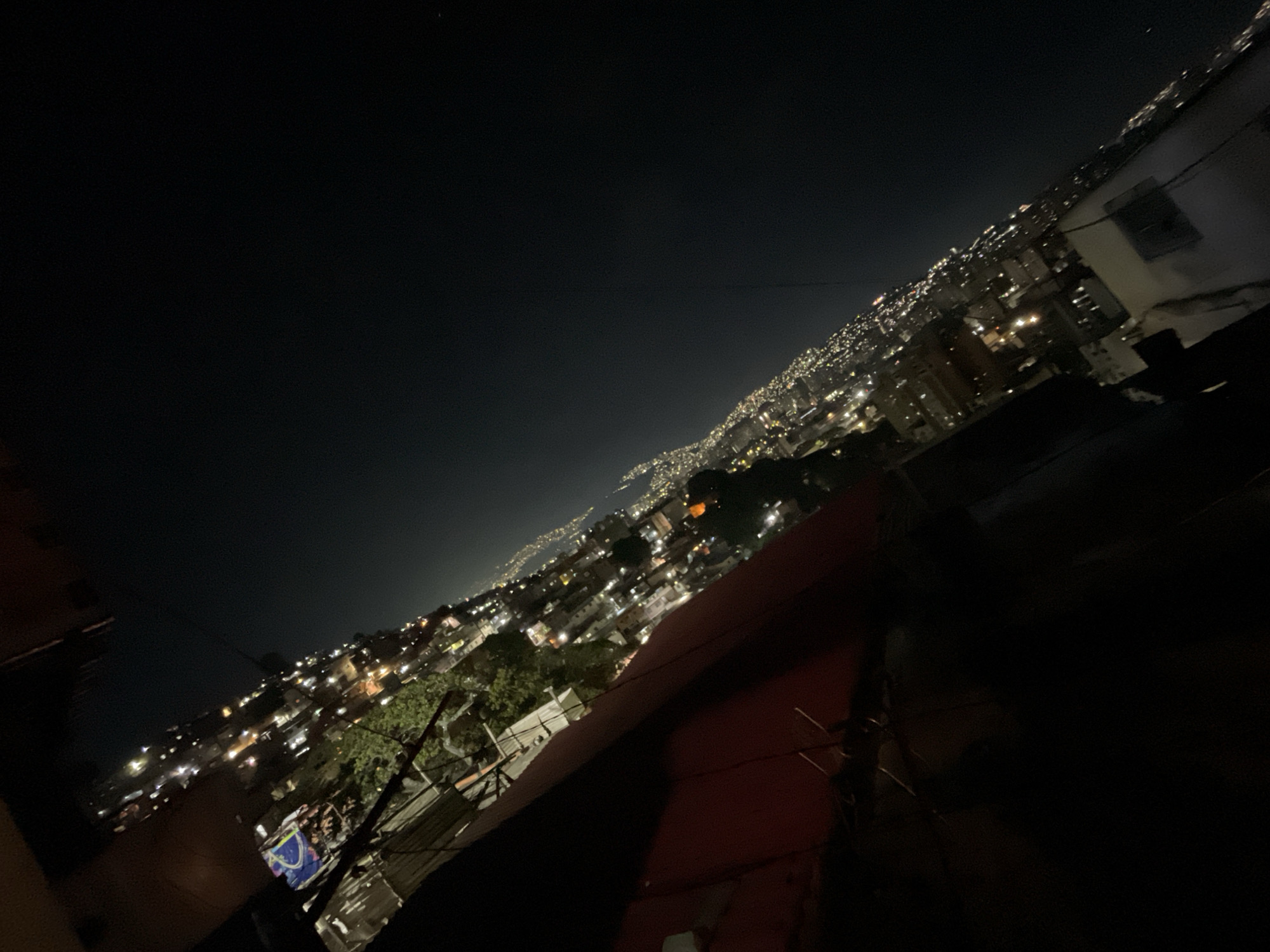A tilted, long-exposure, night view of a city at the base of a hill or mountain range with scattered bright lights, likely from houses and streets. The foreground shows parts of a building or structure with wires and dark areas, giving the impression of looking out from a rooftop. The long exposure blurs the lights and creates light trails suggesting movement, possibly of vehicles. The location is the Petare neighborhood of Caracas, Venezuela.