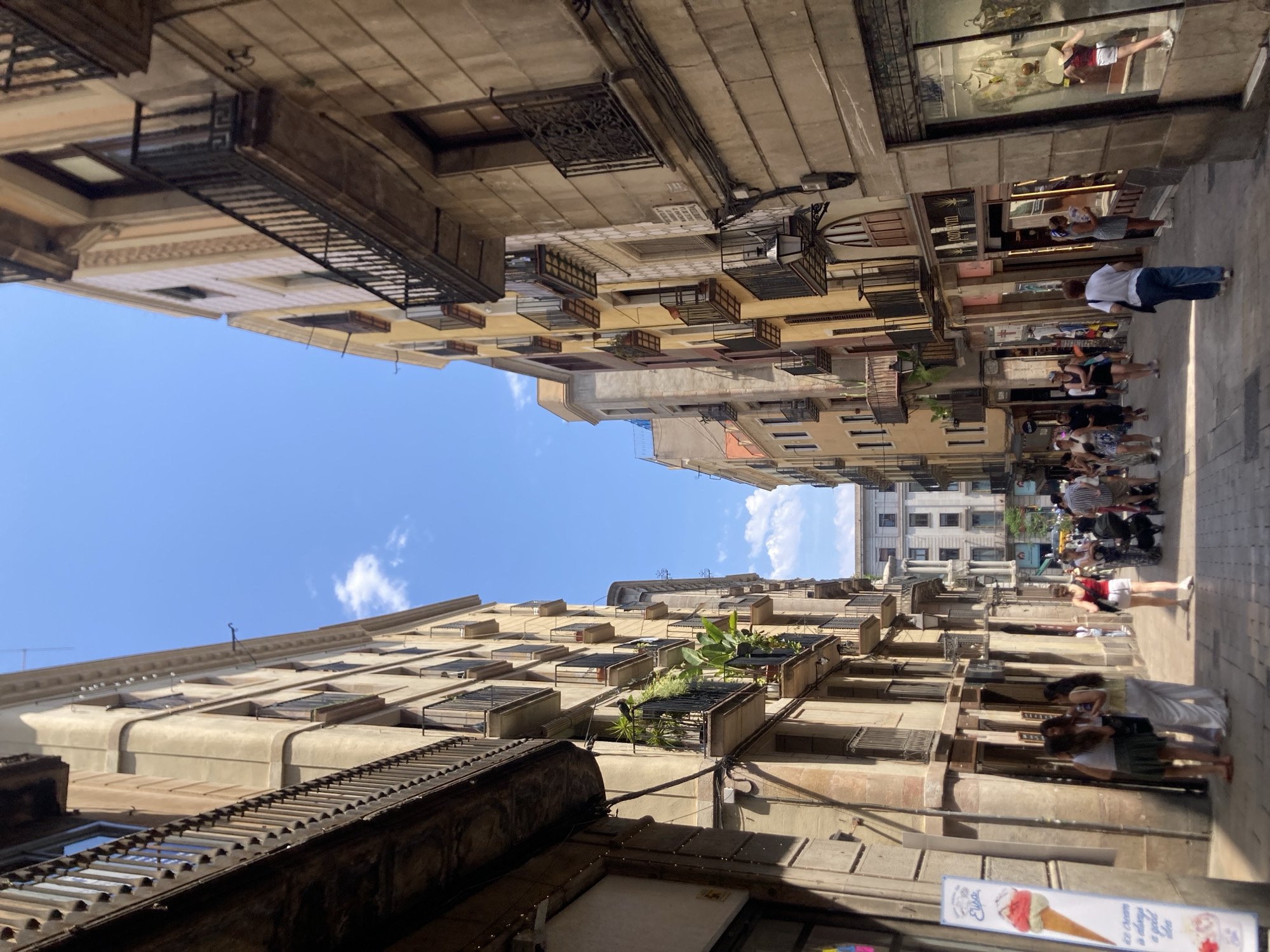 The image depicts a narrow, sunlit street in Barcelona, Spain, lined with multi-story buildings featuring balconies and shops on the ground floor. The architecture is characteristic of older European cities, with stone facades and iron railings on the balconies. A clear blue sky with scattered white clouds is visible above.
On the left side of the street, an ice cream shop display stands prominently, featuring an image of a pink and white ice cream cone and text that reads "ice cream is always a good idea." A group of three women, appearing to be tourists, are standing near the entrance of a shop on the left, looking towards the right. One woman wears a green skirt and white top, another a white top and light-colored pants, and the third a white top and dark skirt.
The street is paved with cobblestones and is populated by pedestrians walking in both directions. A woman in a red top and white shorts is walking away from the camera on the right side of the street, pushing a stroller. Other individuals are seen browsing shop windows and walking along the street. The shops display merchandise, with one window on the right showing clothing on mannequins. The overall atmosphere suggests a vibrant and bustling urban environment during daytime. The lighting indicates it is a sunny day, with strong shadows cast on the street and buildings.