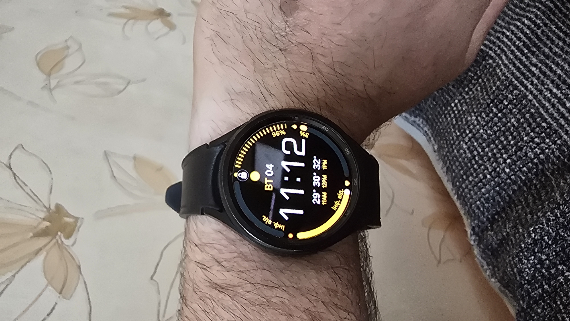 A close-up, overhead shot captures a black smartwatch on a hairy forearm. The smartwatch displays the time as 11:12, with a battery indicator showing 96% and a small droplet icon. Below the time, it shows three temperature readings: 29°, 30°, and 32°, with corresponding times of 11 AM, 12 PM, and 1 PM. The watch face also features the text "BT 04" and some Ukrainian text. The background is a light-colored wallpaper with a floral pattern. The image does not provide any information to determine the location in the city of Novohuivynske, Ukraine.