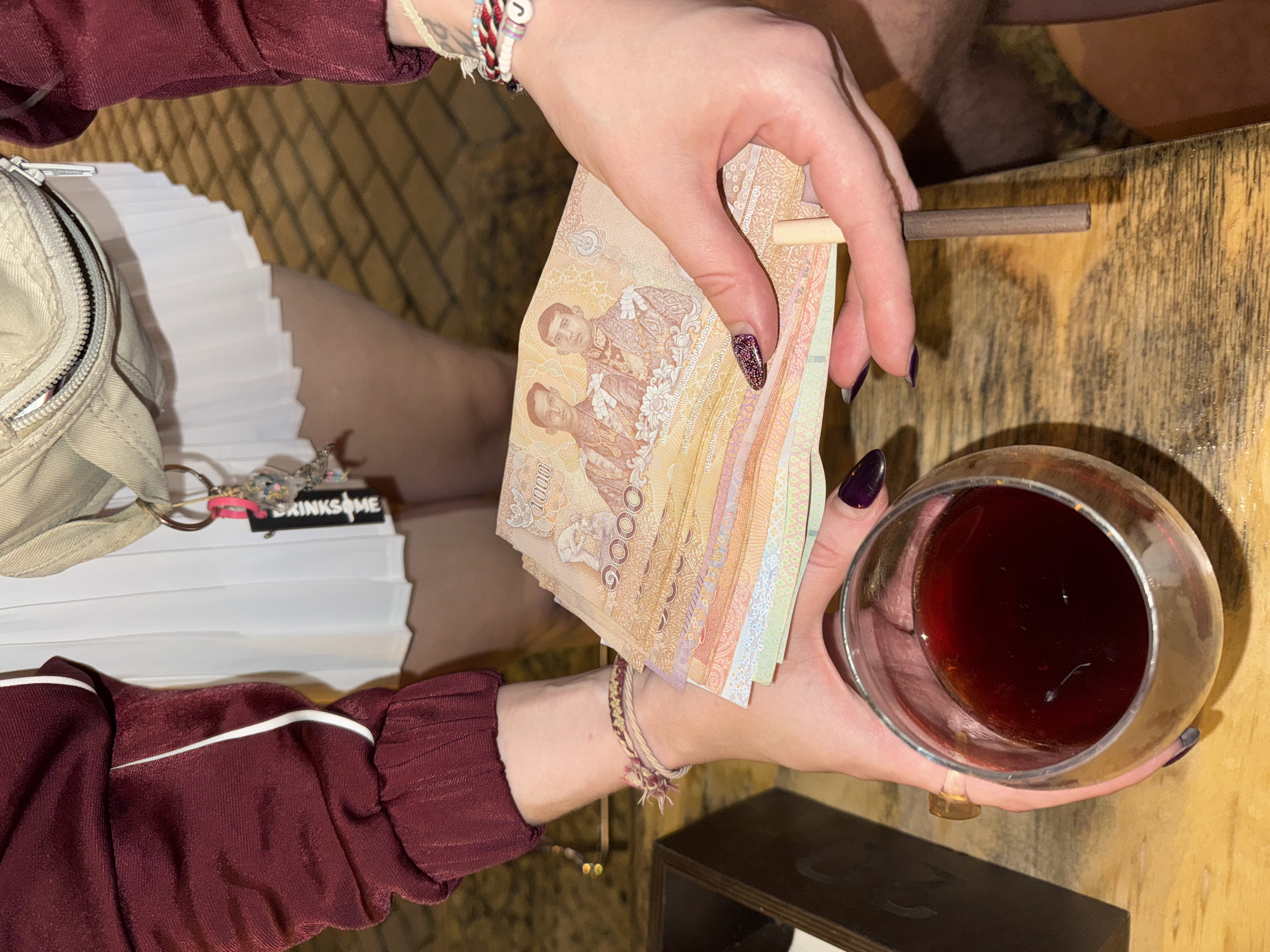 In an indoor environment, a close-up shot focuses on a person's hands holding a stack of Thai baht banknotes and a glass of dark red liquid. The banknotes are predominantly orange and yellow, with images of two men in regal attire visible. The person's hands, with dark purple nail polish, are carefully holding the money, which is fanned out. One hand also holds the stem of the glass, which has a golden rim and contains a dark, possibly alcoholic, beverage. The background is a blurred wooden surface. The person is wearing a maroon jacket with white trim and a white pleated skirt. A light-colored fanny pack with a zipper is visible around the waist. The overall lighting suggests an evening or indoor setting, possibly a bar or restaurant. The image is taken from a slightly elevated angle, looking down at the hands and the items they hold.