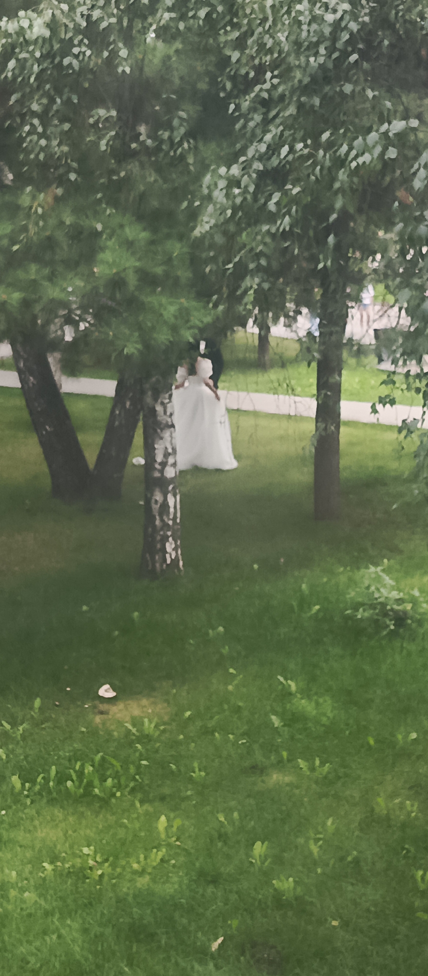The image shows a wedding scene outdoors, possibly in a park or garden. A bride in a white wedding dress is visible, with her back to the camera, and a person in a dark suit, presumably the groom, is standing behind her. They are on a green lawn with trees surrounding them. A paved path runs through the background, and there are some buildings or structures in the distance. The lighting suggests it is daytime, and the atmosphere is somewhat diffused, possibly due to overcast skies or foliage. There are a few fallen leaves or small objects on the grass. The overall impression is a quiet, intimate moment during a wedding celebration.