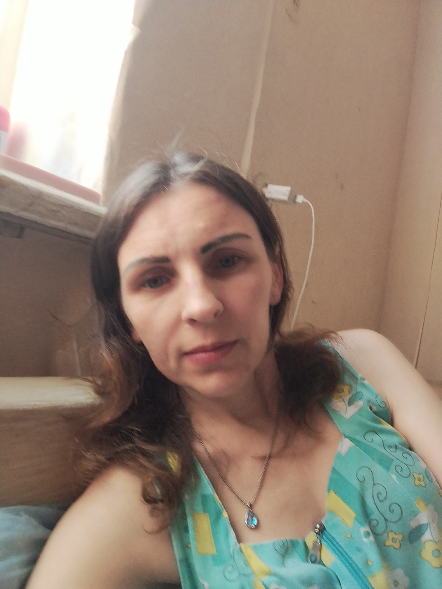 A woman with dark brown hair and blue eyes is posing for a selfie. She is wearing a turquoise and yellow floral print dress with a zipper down the front. A silver necklace with a blue pendant is visible around her neck. She is indoors, with a window and a wall visible in the background. There is also a white electrical cord and plug on the wall. The scene appears to be in a residential setting. There is no information about the location within the city of Vinnytsia, Ukraine.