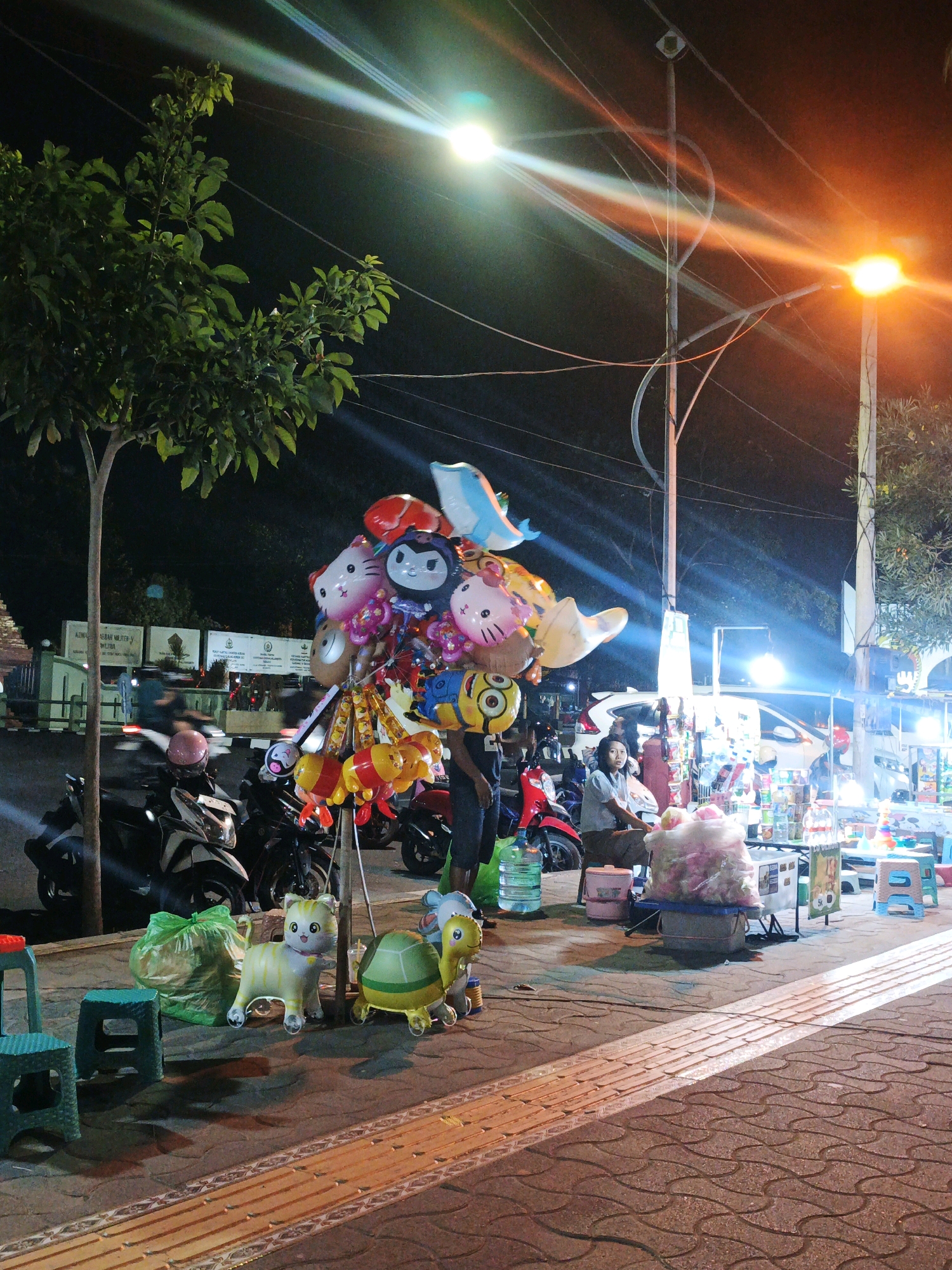 The image depicts a nighttime street scene in Kauman, Indonesia, likely a market or roadside stall area. A vendor is seated behind a stall laden with various goods, possibly snacks or small toys, illuminated by bright overhead lights. In the foreground, a collection of character balloons, including Hello Kitty, Minions, and other cartoon figures, are displayed, attached to a stand. To the left of the balloons, there are several small plastic stools and a large green garbage bag. Further left, a tree with dense green foliage stands tall. Several motorcycles are parked behind the tree and balloons, suggesting a busy thoroughfare or gathering spot. The ground is paved with interlocking stones. The overall atmosphere is lively, with colorful lights and illuminated goods creating a festive ambiance.