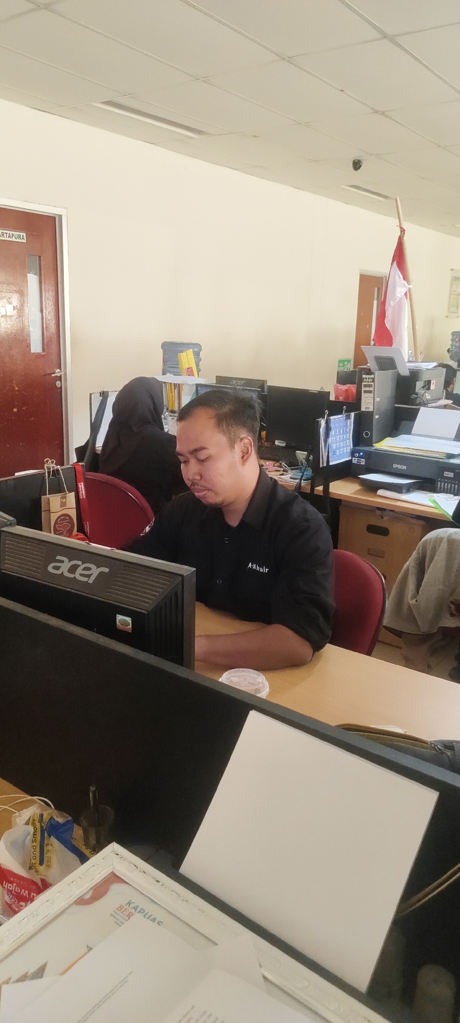 The image depicts an indoor office environment. A man in a black collared shirt is seated at a desk, looking down at a computer monitor. The shirt has a white embroidered logo on the right side of the chest that reads "A-Khair". He appears to be focused on his work. In the background, a person wearing a black hijab is visible, also seated at a desk. The office contains multiple computer monitors, a printer, filing cabinets, and a water cooler. A red and white Indonesian flag is displayed on a pole in the background. Various office supplies and personal items are scattered on the desks. The overall setting suggests a typical office workspace in Nusa Indah, Indonesia, possibly during work hours.