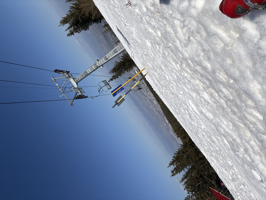 The image displays a snow-covered ski slope on a clear, sunny day, providing a panoramic view of a distant city situated in the valley below, consistent with the area near Bistritsa, Bulgaria. The immediate foreground, in the lower right, shows the partial toe of a red ski boot. The snow on the slope is uneven and textured. A tall metal ski lift tower, featuring multiple cables and two unoccupied chairlift seats, dominates the upper right-center of the frame, extending upwards. On the slope to the left of the tower, a blue sign with white lettering or a logo is attached to a yellow marker pole, alongside another yellow pole and a black rectangular sign. Dark green evergreen trees line both the left and right edges of the ski slope. The background reveals a wide, clear blue sky over a sprawling urban landscape in the distant valley, with subtle haze and faint mountain ranges on the horizon. A red tarp or awning, with an indistinct, obscured figure beneath it, is faintly discernible in the far lower-left quadrant of the image.