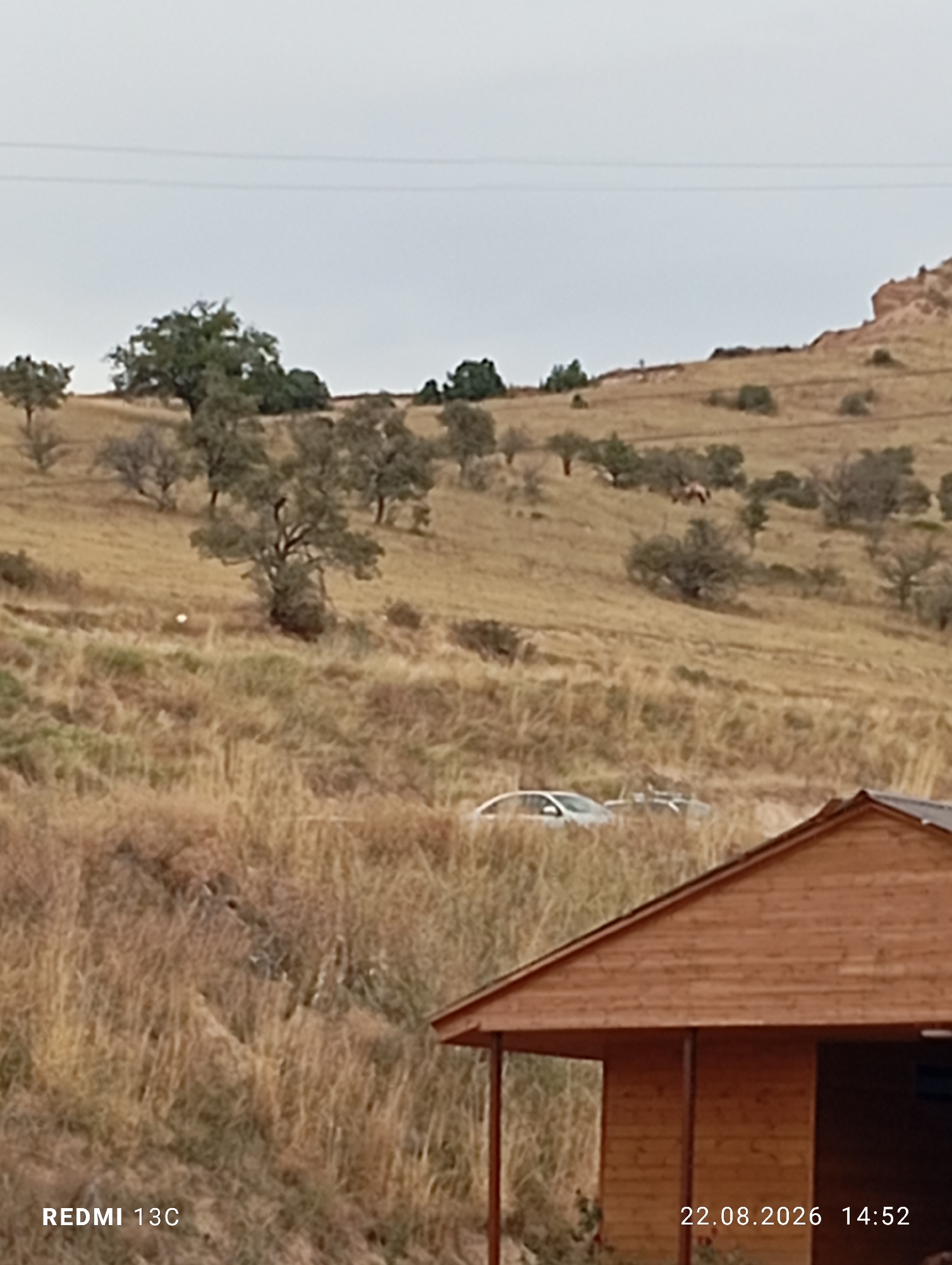 The image shows a rural landscape with rolling hills covered in dry grass and scattered trees. A few cars are parked on a dirt road on the hillside. In the foreground, a wooden structure with a pitched roof is visible. The timestamp at the bottom right indicates the date and time as 22.08.2026 14:52. The text "REDMI 13C" is present at the bottom left, indicating the device used to capture the image. There are no people visible in the image. The overall scene depicts a natural environment with evidence of human activity. This scene is likely located in the vicinity of Amirsay, Uzbekistan, known for its natural beauty and mountainous terrain.