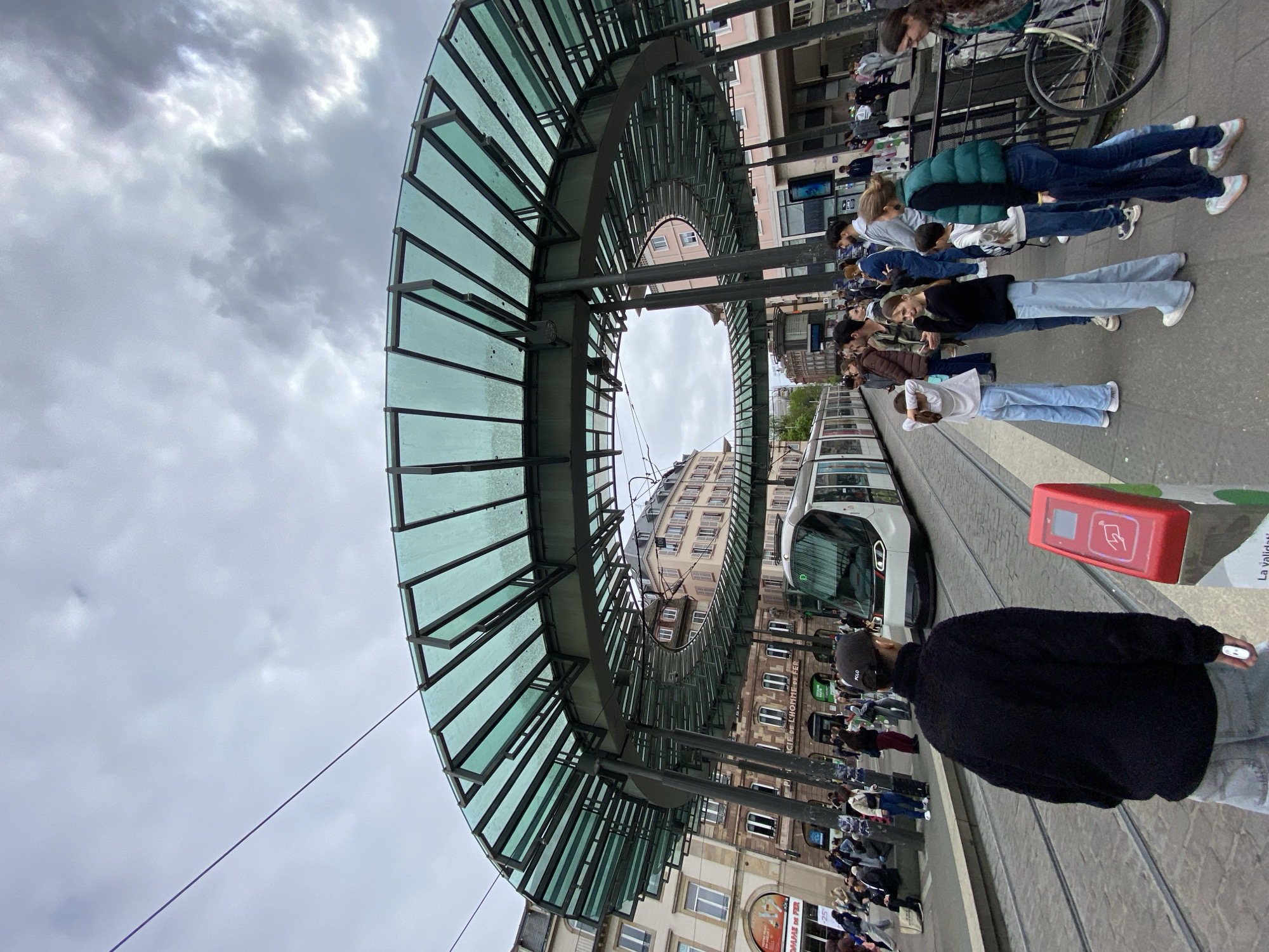 The image shows a street scene in Strasbourg, France, likely near a tram stop. The architecture features a curved, glass-covered structure over the tram tracks. A modern tram is visible on the tracks. People are standing on the sidewalk, appearing to wait for the tram. The sky is overcast. The buildings in the background are typical of European urban architecture. A fare validation machine can be seen near the tram tracks. The environment suggests a public transportation hub in an urban setting.