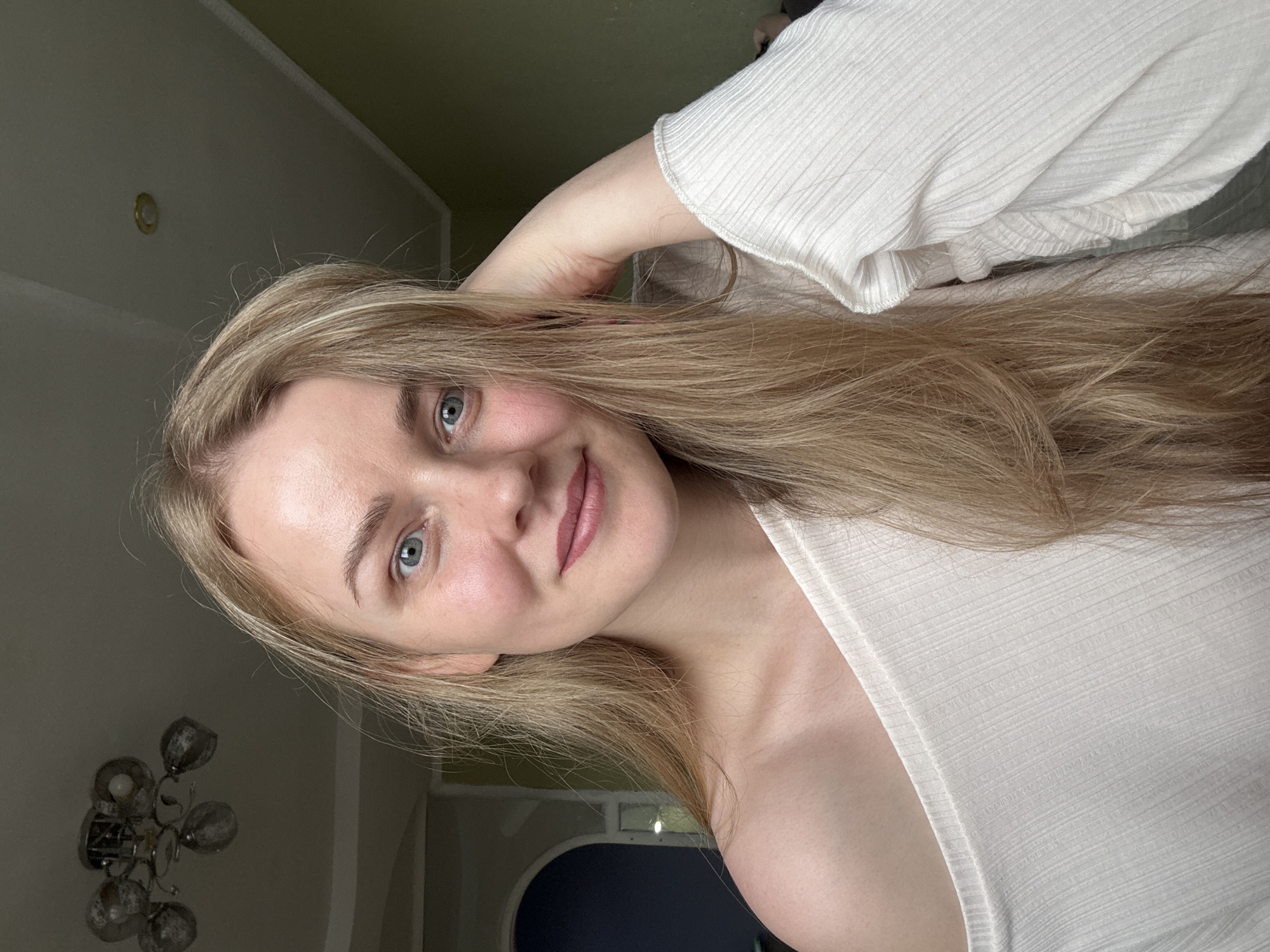 A woman with long blonde hair is looking at the camera. She is wearing a white top and has one hand behind her head. A chandelier is visible in the background. It is unlikely that the woman in the photo is in Chaikovsky, Russia.