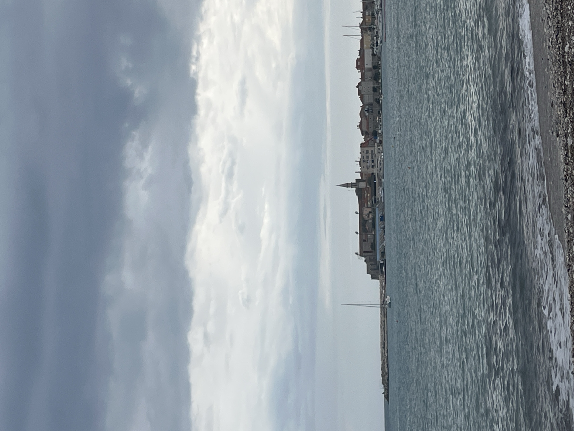 The image shows a coastal view of Budva, Montenegro, under an overcast sky. The scene is divided between the sky with heavy, gray clouds and the Adriatic Sea. The sea, marked by ripples, stretches to a pebble beach. Along the coastline, buildings, including a prominent church steeple, indicate the urban layout. A few boats are visible in the water, with one in particular closest to the camera, standing out as a clearer subject. The interaction between the sea and the coastline creates waves washing onto the shore.