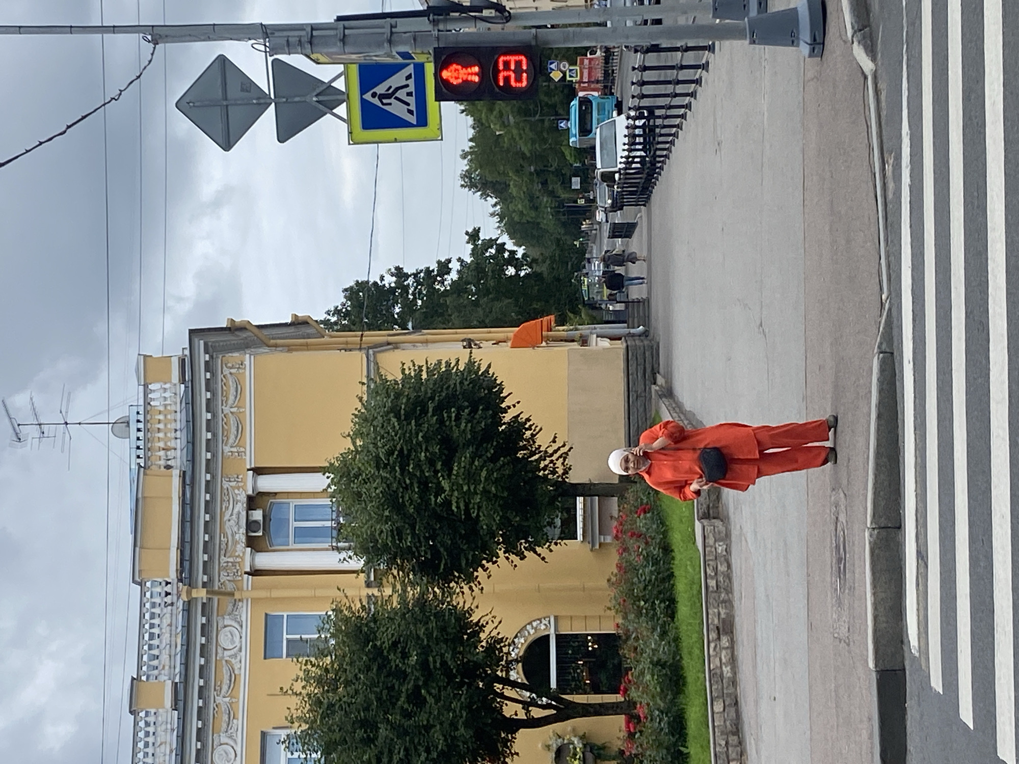 The image depicts a woman standing on a crosswalk in Pushkin, Russia. She is wearing an orange headscarf and an orange pantsuit, and she is holding a black bag and talking on a mobile phone. The woman is positioned in the foreground, facing the viewer. Behind her, a yellow building with ornate architectural details stands prominently. Trees and flowerbeds are visible in front of the building. The background features a sidewalk with pedestrians, traffic signals displaying a red pedestrian icon and the number 23, and vehicles on a street. The sky is overcast. The scene appears to be a street in a European city, likely Pushkin, given the historical architecture.