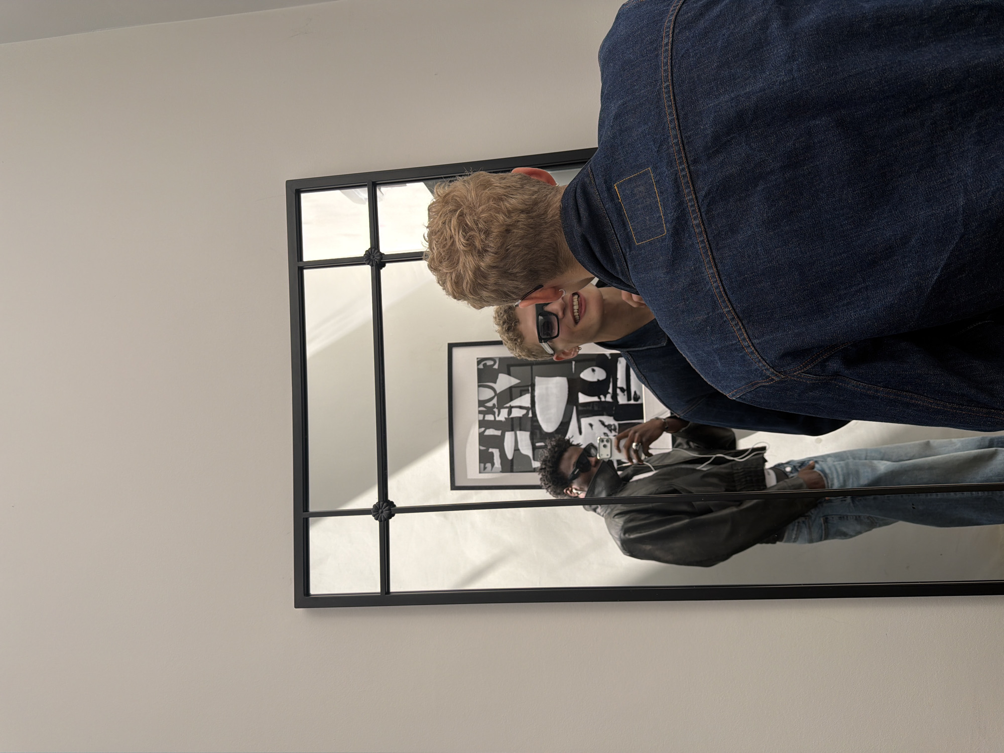 A medium shot captures two young men from behind and reflected in a large, multi-paneled mirror. The man in the foreground, facing away from the camera, has short, light brown curly hair and is wearing a dark blue denim jacket. In the mirror's reflection, the same man is seen from the front, smiling and wearing sunglasses. To his left, a second man, reflected in the mirror, has dark skin, curly dark hair, sunglasses, a black mask covering the lower half of his face, and is wearing a dark jacket over a white shirt and blue jeans, holding a phone. He is taking a selfie in the mirror. A framed artwork with black and white graphics is visible on the wall behind the reflected figures. The scene is set against a plain, light-colored wall, suggesting an indoor environment, possibly a home or studio, with no discernible London landmarks.