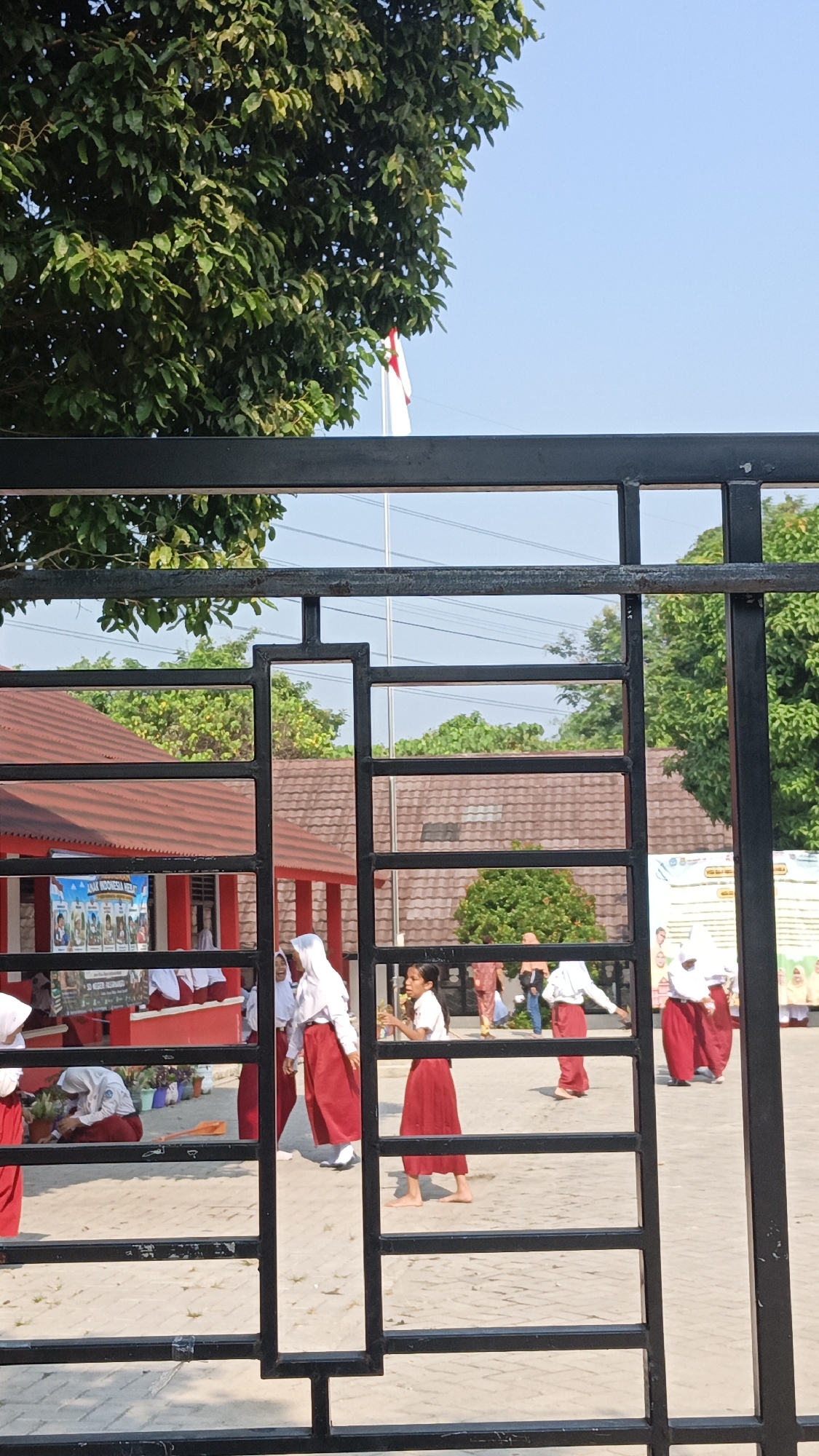 A high-angle, outdoor, medium shot captures a group of students, appearing to be elementary school-aged, in a courtyard with a building and trees in the background. The students are dressed in white shirts and red skirts, typical school uniforms in Indonesia. Several students are engaged in various activities; one student is planting in a pot, another is walking with a flower, and others appear to be socializing. The courtyard is paved with interlocking bricks. A black metal gate with a geometric design partially obscures the view, with a glimpse of a red-roofed building with an outdoor walkway. In the background, a tall flagpole with a red and white flag, the flag of Indonesia, is visible against a clear blue sky. The overall impression is a sunny day at a school in Pasir Bolang, Indonesia, with students participating in outdoor activities.