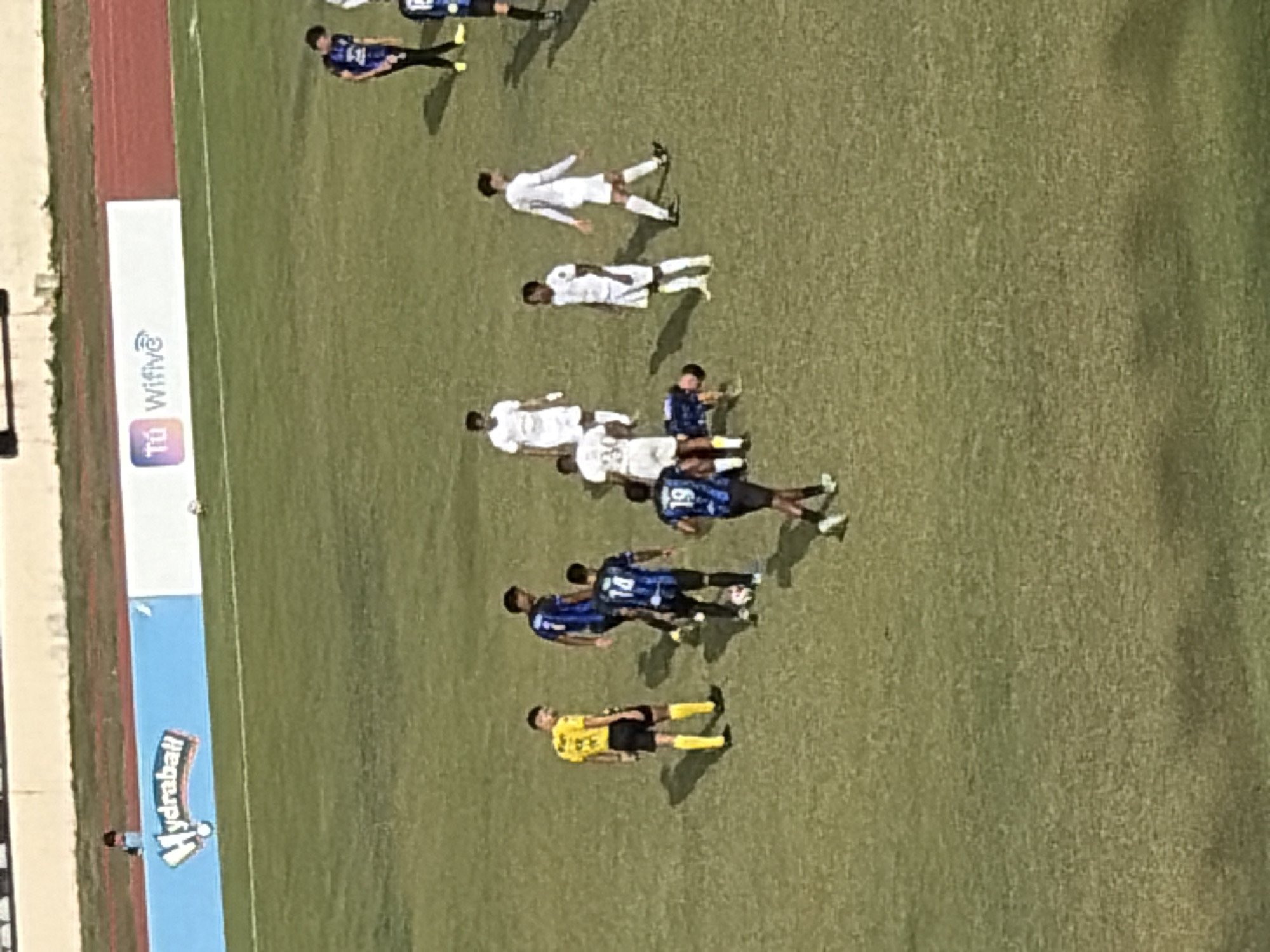 The image depicts a soccer field during what appears to be a match. The field is bordered by a running track and an advertising banner. Multiple players are visible on the field, wearing either white or blue uniforms. One individual is wearing a yellow referee uniform. The players seem to be in various positions. The match is being held in Ciudad Guayana, Venezuela.