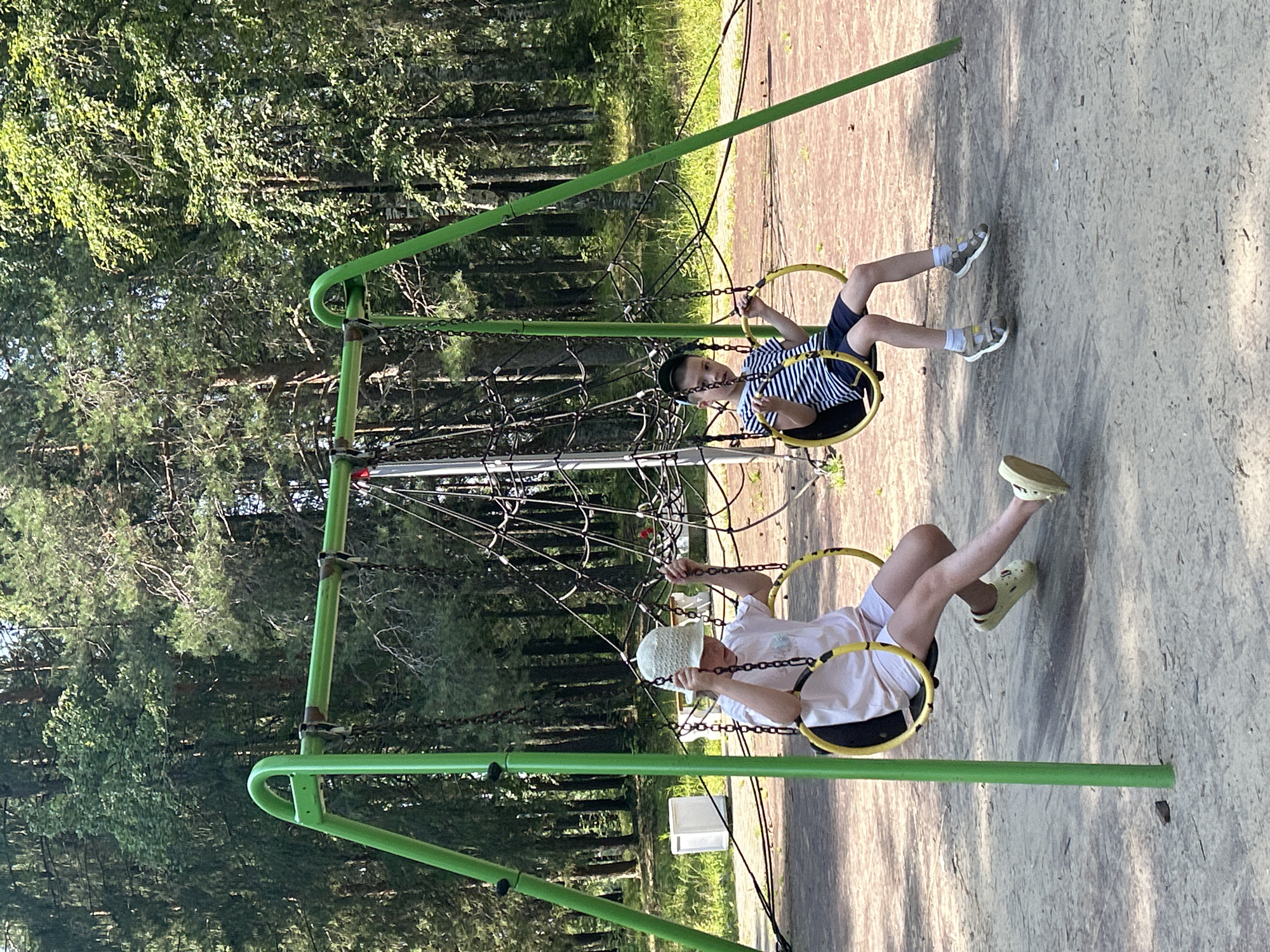 In Krasnovishersk, Russia, a public playground features two children on swings. On the left, a child wearing a light-colored hat and t-shirt is seated on a yellow-ring swing, their legs extended forward. To the right, another child, wearing a striped t-shirt and dark shorts, sits on a similar swing, with their legs bent. Both children are holding onto the swing chains. The swings are suspended from a green metal A-frame structure. Behind the children, a rope climbing net is visible. The ground beneath the swings is a mix of sand and reddish-brown material. The background consists of a dense forest with tall pine and birch trees, illuminated by sunlight filtering through the leaves. A white rectangular object, possibly a trash bin or a utility box, is situated in the wooded area to the left. The scene depicts a typical summer day at a park.