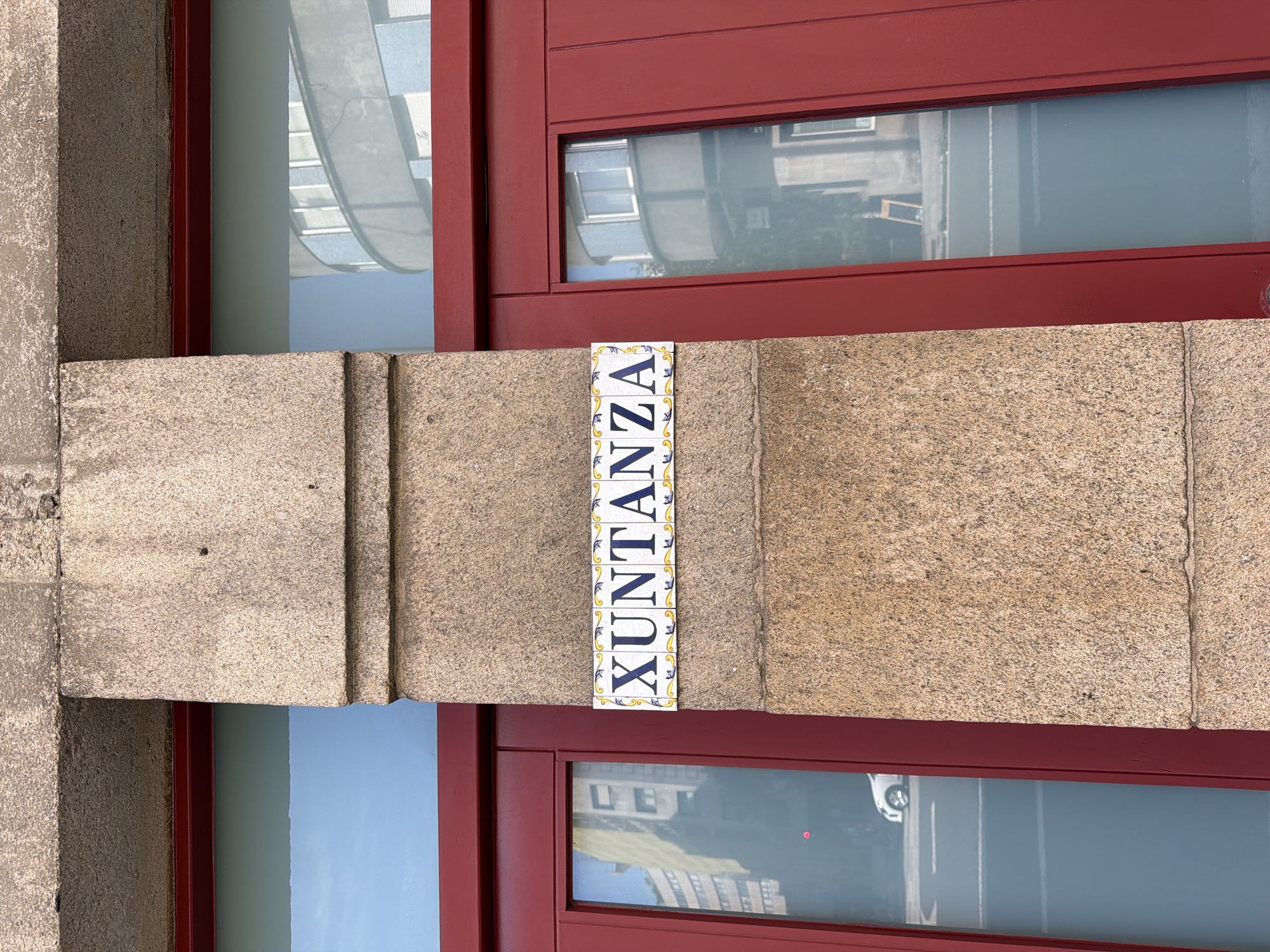A close-up shot captures a decorative tile sign with the name "XUNTANZA" against a rough, granite column. The sign features blue lettering with yellow floral accents on a white background. The column is flanked by deep red double doors, with a large glass window to the left and a segmented glass panel on the right. Reflections in the windows show elements of the surrounding urban environment, including buildings, a street, and a white car. The upper part of the image reveals the textured stonework of a building facade. The lighting suggests it is daytime, casting natural light on the scene. The location is identified as Santiago De Compostela, Spain.
