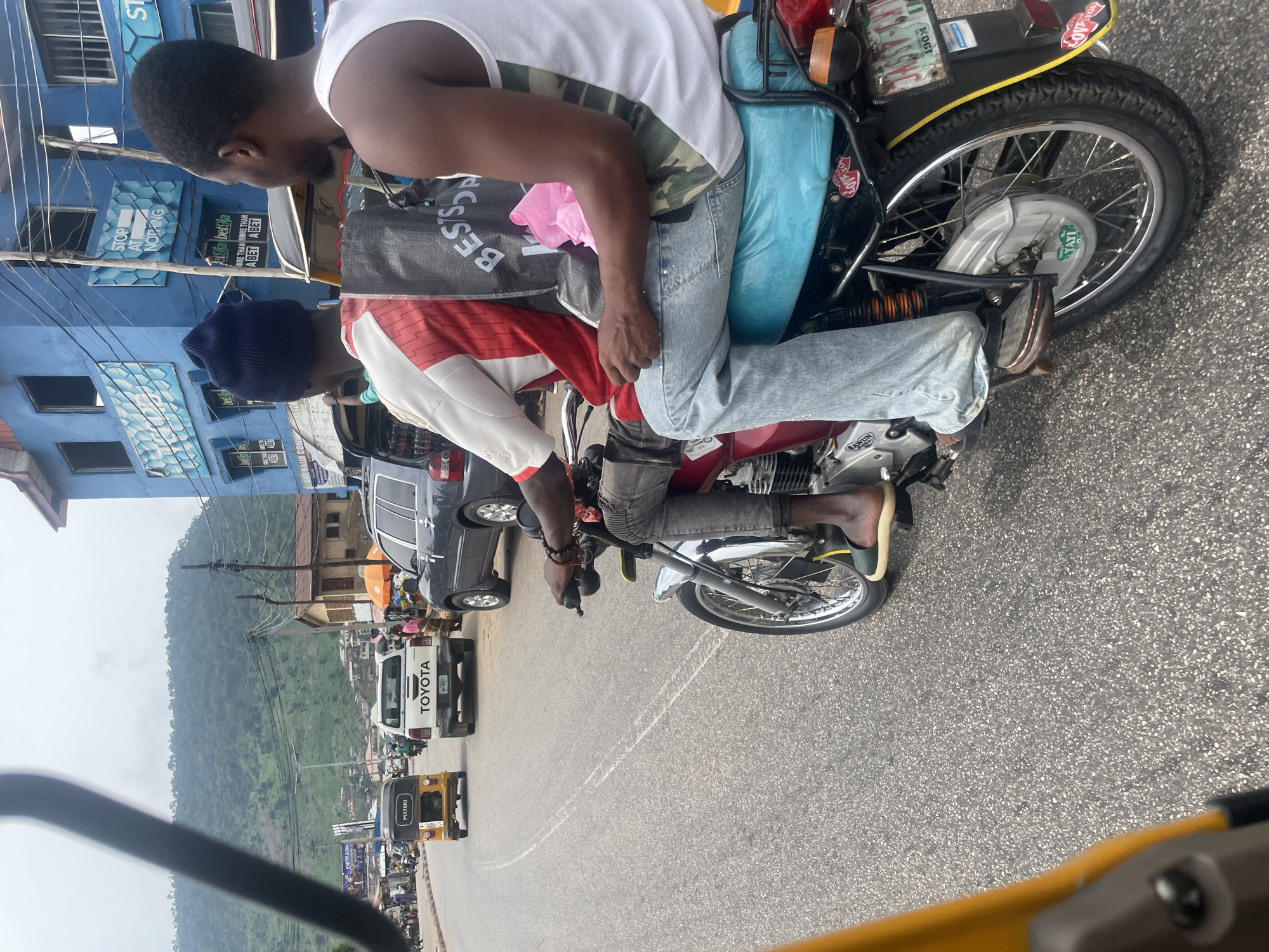 Two men are riding on a motorcycle on a paved road in Lokoja, Nigeria. The man in the front is wearing a blue beanie, a red and white shirt, and blue jeans. The man in the back is wearing a white tank top with a camouflage pattern. He is holding a black bag with white lettering that says "BEST SHOP". The motorcycle is red and has a blue seat. A pink plastic bag is tucked under the man in the back's arm.
In the background, there are several vehicles, including a yellow tricycle, a white Toyota pickup truck, and a dark-colored SUV. Buildings line the street, one of which is painted blue and has signs on it. Hills covered in vegetation are visible in the distance. The sky is overcast. The overall scene depicts a typical street view in Lokoja, Nigeria, with various modes of transportation and commercial activity.