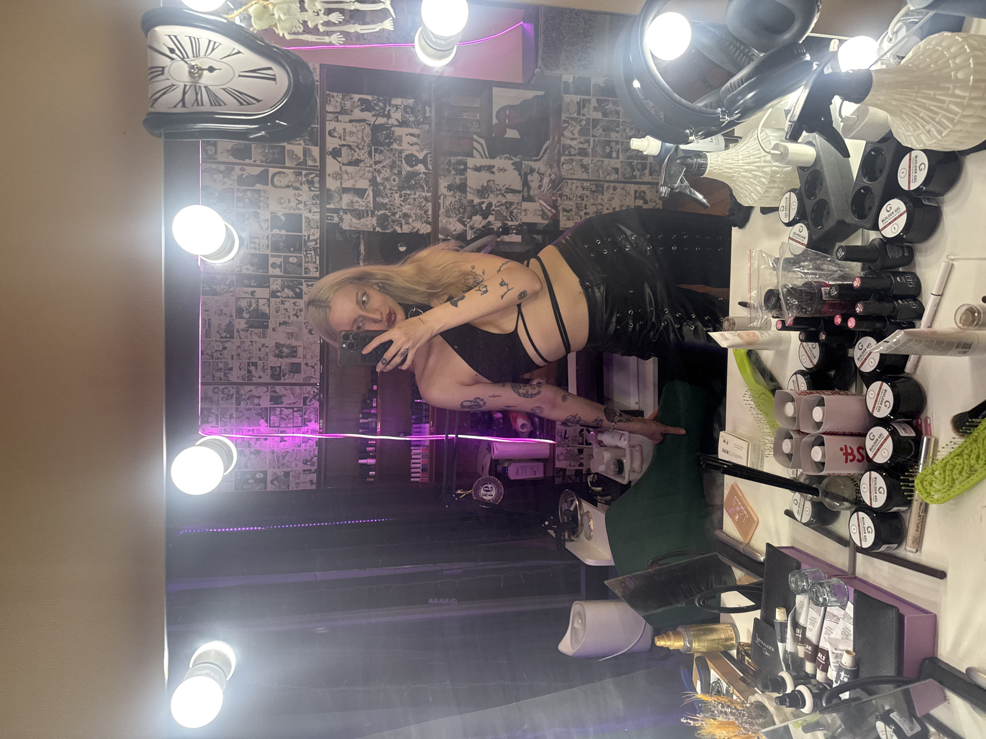 The image depicts a woman taking a mirror selfie in what appears to be a dressing room or vanity area. The woman has blonde hair, is wearing a black top and black leather pants, and has numerous tattoos on her arms. She is holding a smartphone and is posing with one hand on her hip.
The background features a large mirror illuminated by vanity lights. The wall behind the mirror is decorated with what looks like a collage of black and white manga or comic book pages. To the right, a novelty clock in the style of Salvador Dalí's "The Persistence of Memory" is visible, along with a small skeleton figure.
On the vanity table in front of the mirror, there are various cosmetic and nail products, including bottles, jars, brushes, and tubes. A green chair is partially visible on the left side of the image, and a white humidifier is also present. The overall lighting is dim, with purple and white lights from the vanity and other sources creating an atmospheric effect.
There is no information in the image to suggest that the location is Moscow, Russia.