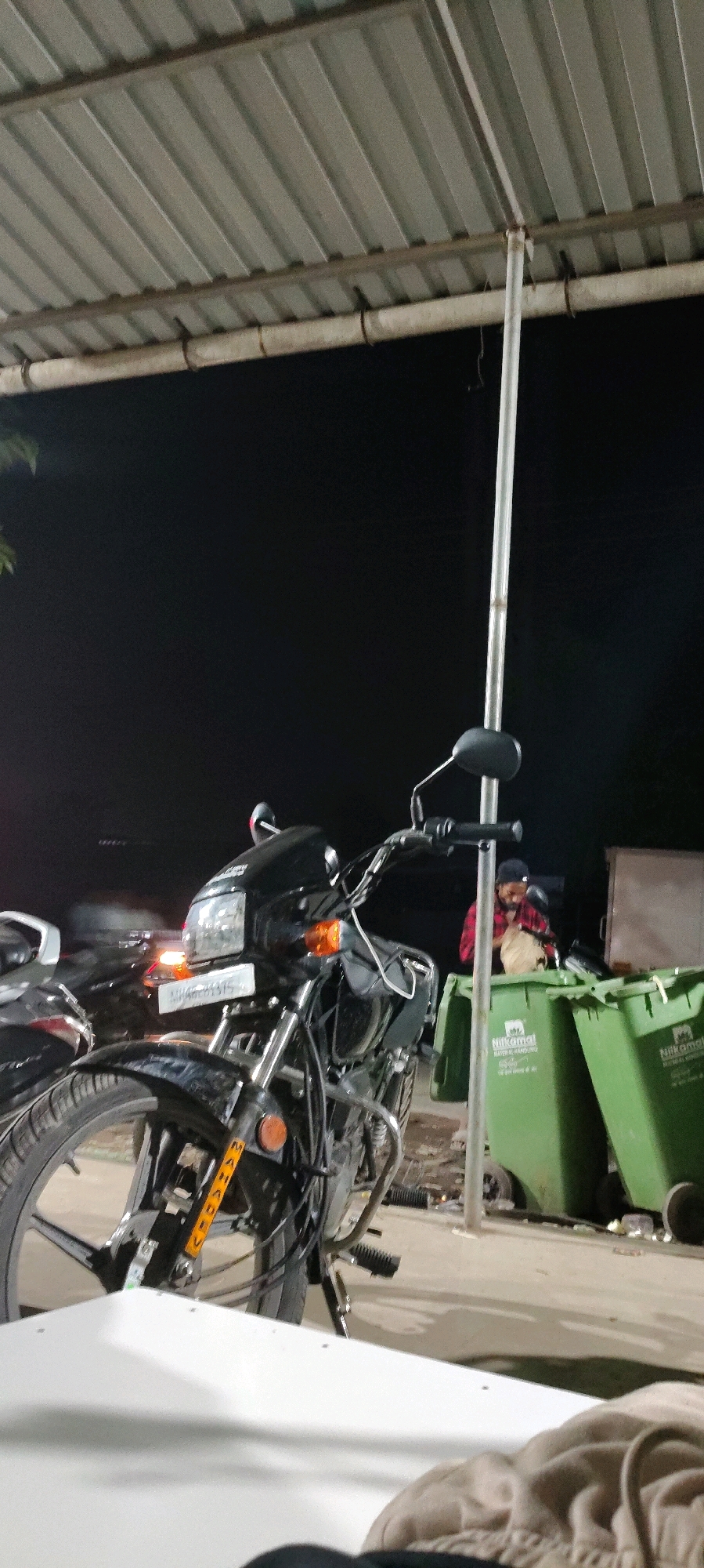 In Vasai-Virar, India, at night, a black motorcycle is parked under a metal roof supported by a pole. A person wearing a red and black checkered shirt and a black cap is standing next to two green dumpsters, sorting through a bag. The dumpsters have text printed on them, likely indicating a municipality or service provider. In the background, another motorcycle is partially visible. The scene appears to be outdoors, possibly at a roadside stall or a public area. The lighting is dim, suggesting it is after sunset.